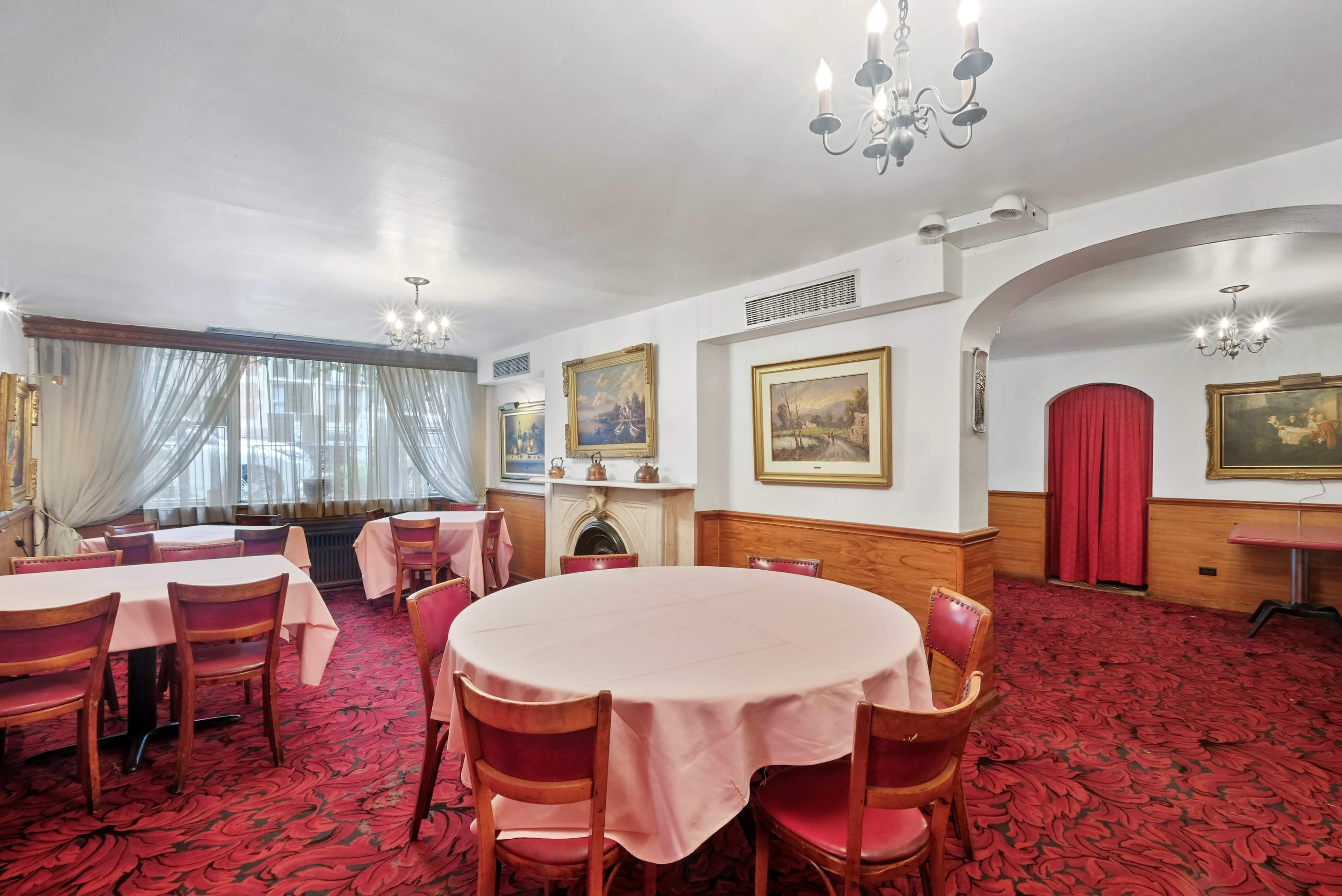 An elegantly furnished dining room with round tables covered in pink tablecloths, red patterned carpeting, and framed paintings on the walls.