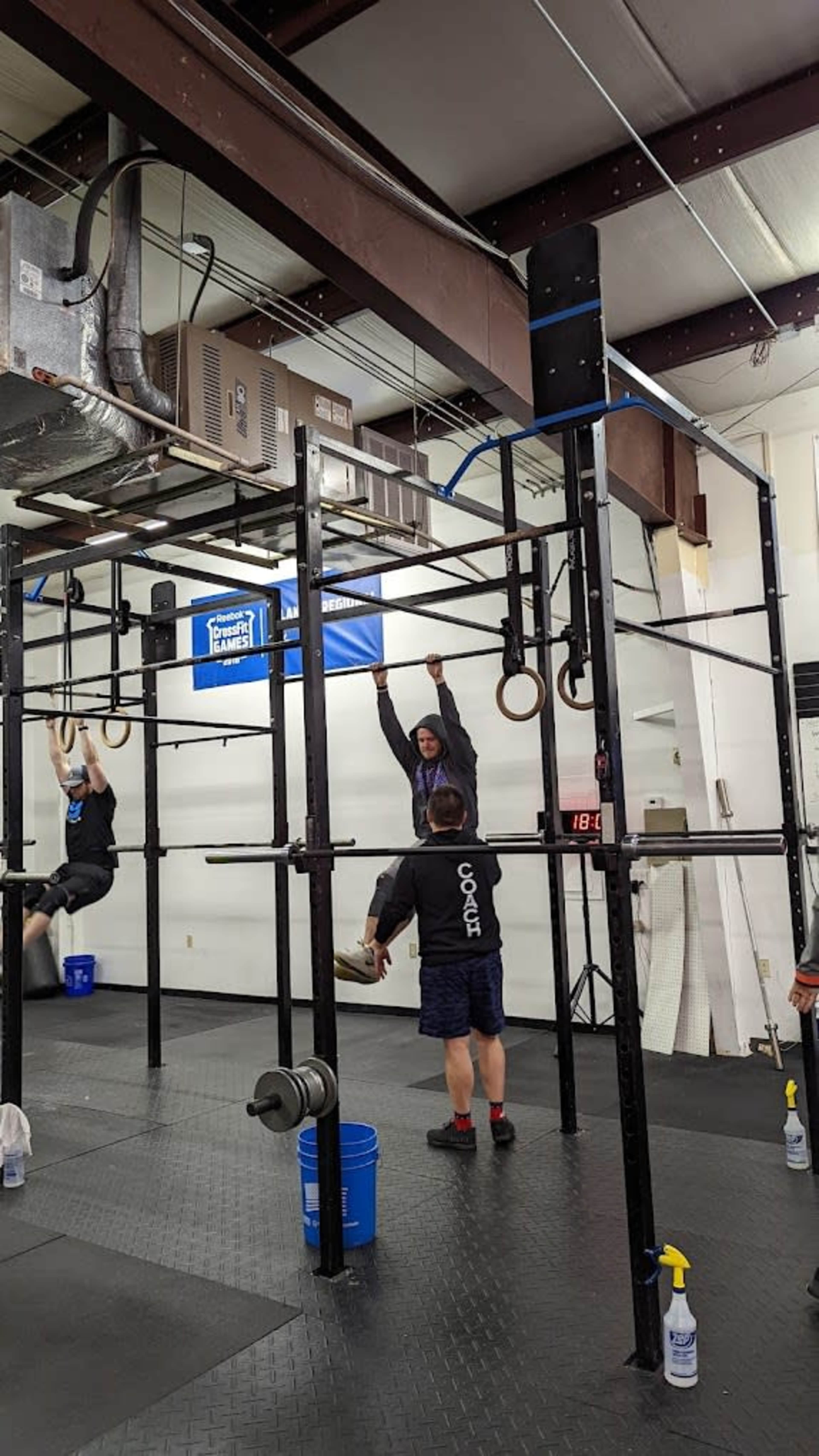 A person is hanging from gymnastic rings in a fitness facility, while another individual performs a strength exercise on the pull-up bar nearby.