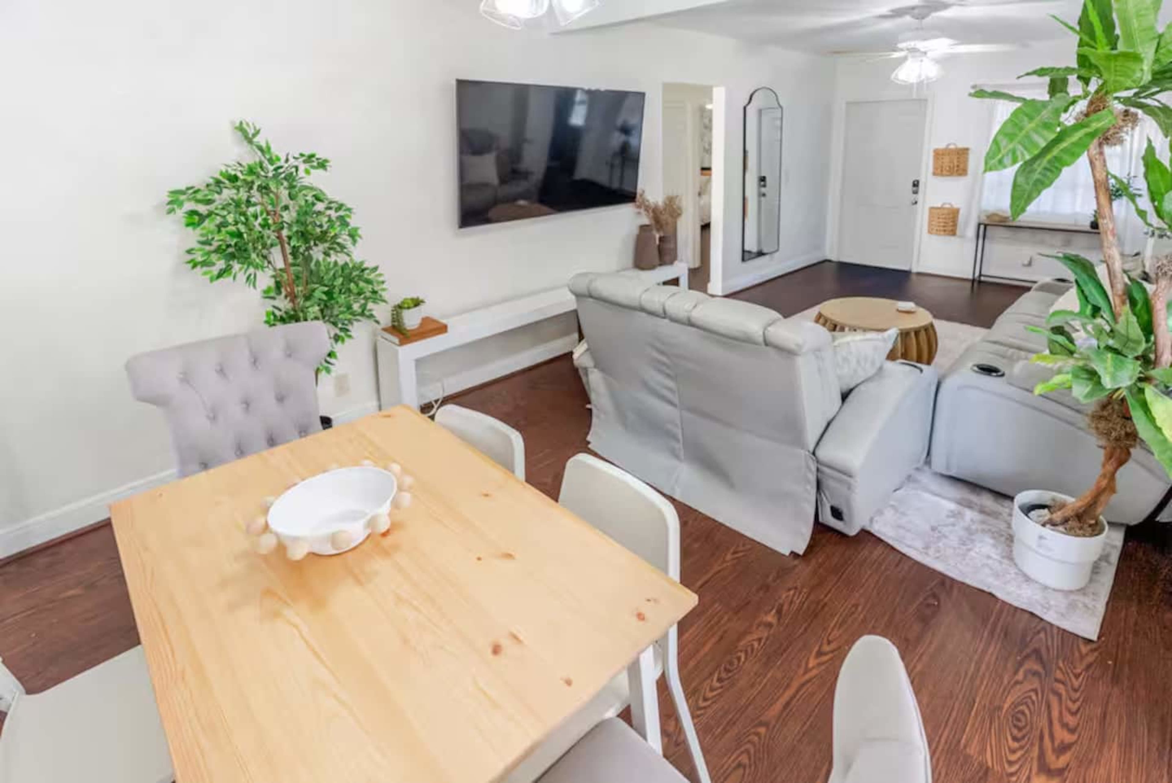 The image shows a bright living space with a dining table, a sectional sofa, a television mounted on the wall, and various indoor plants.