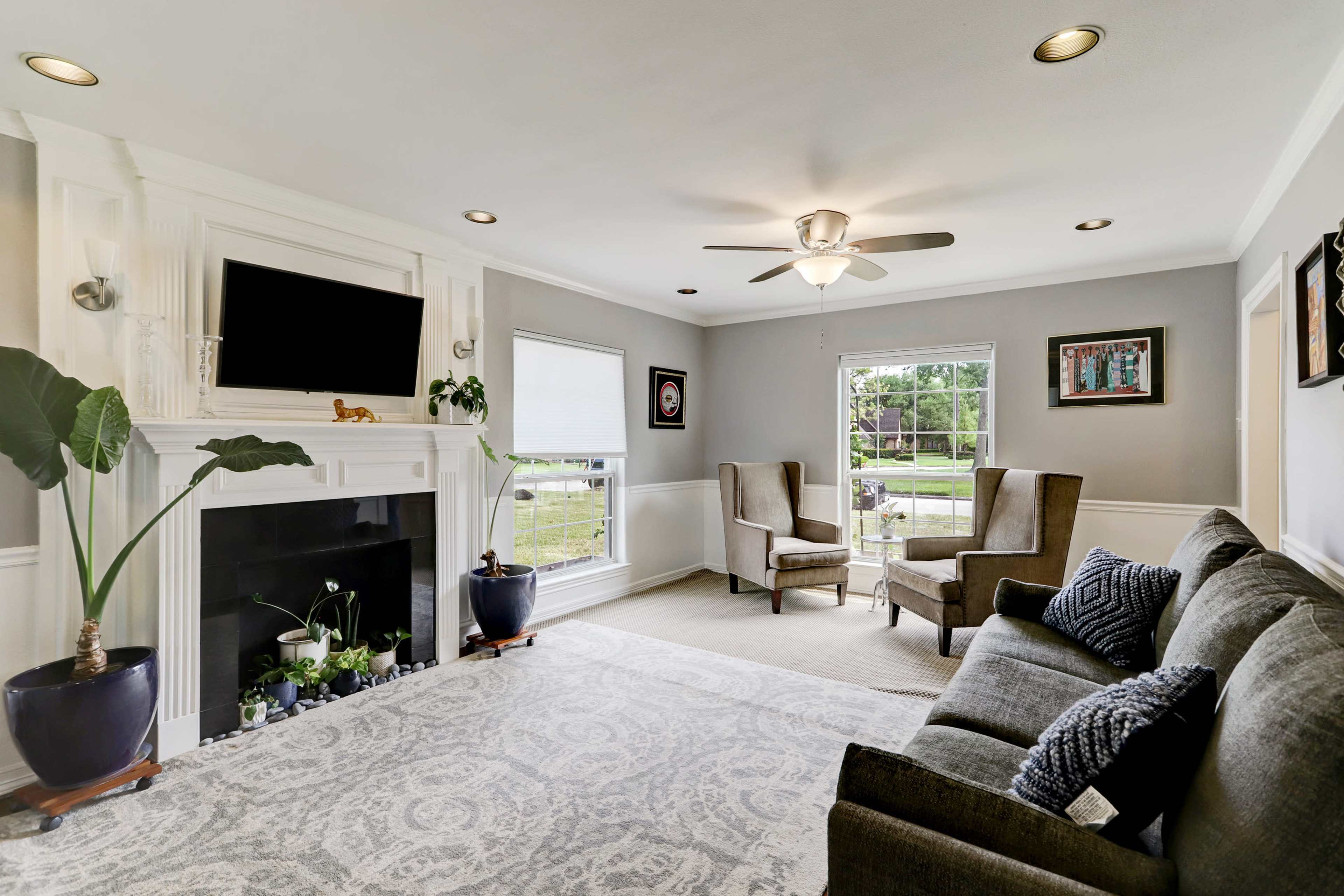 The living room features a pair of armchairs facing a couch, a fireplace with a television above it, and large windows allowing natural light to flood the space.