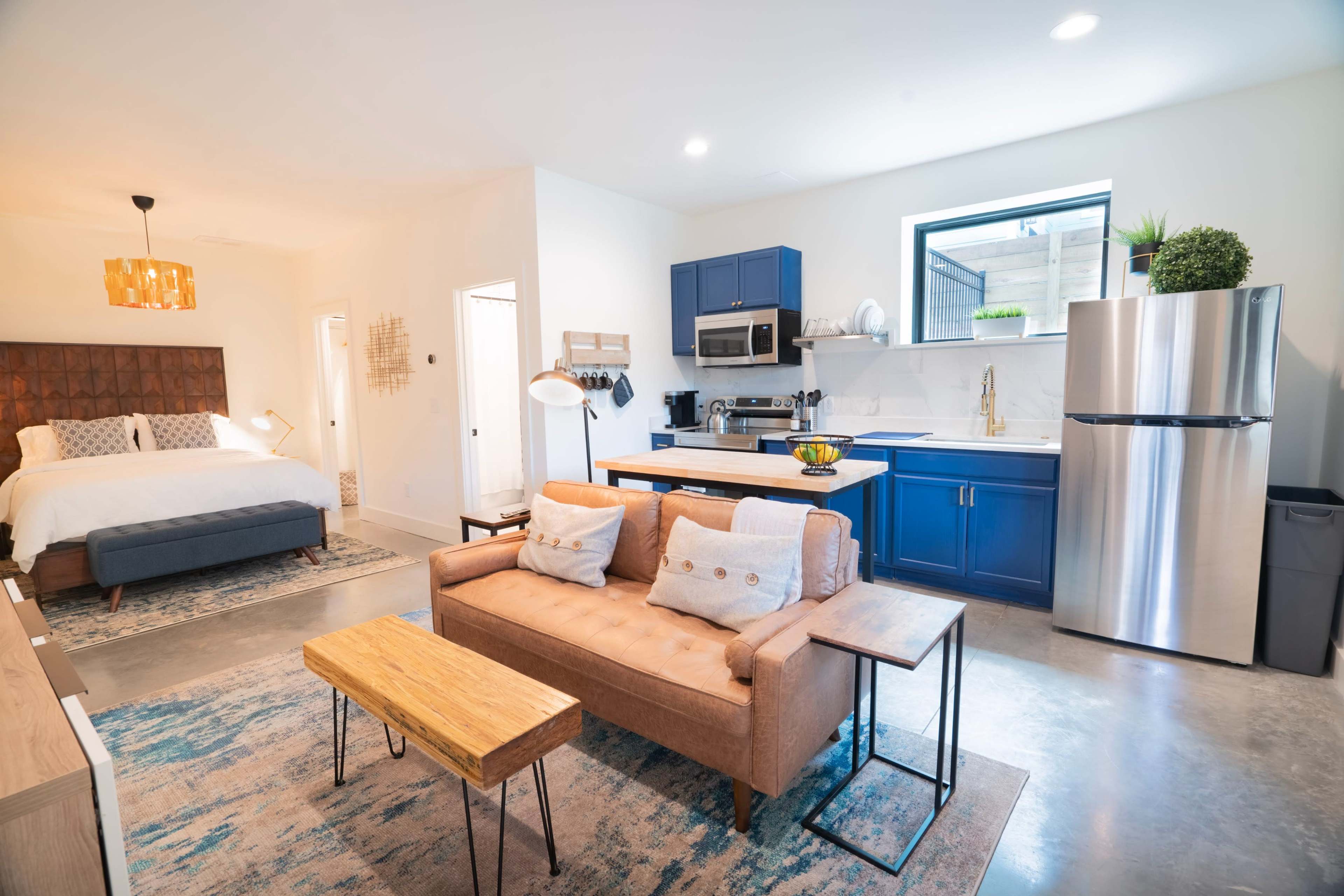 A modern, open-concept living space featuring a bedroom area with a bed, a kitchen with blue cabinetry, and a seating area with a sofa and coffee table.