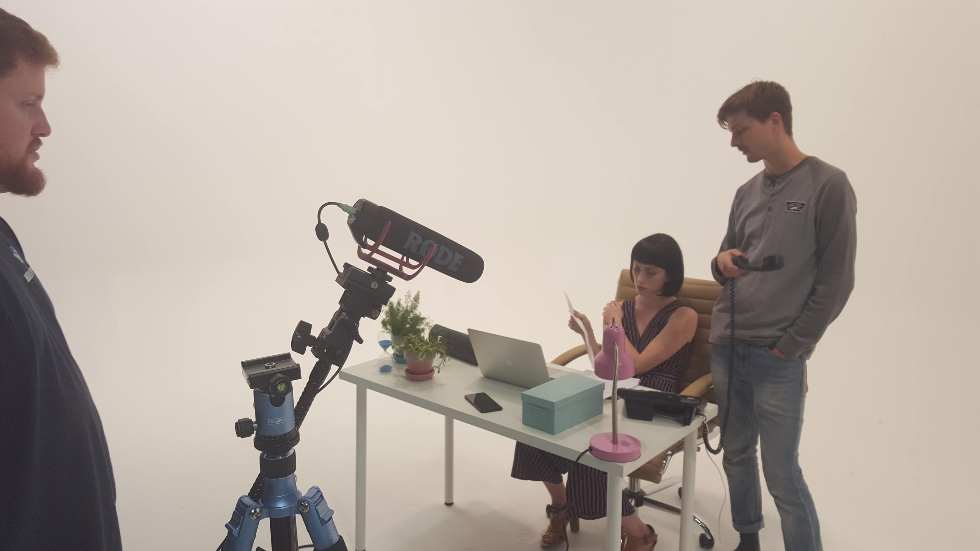 A woman sits at a desk with a laptop and props, while two men adjust camera equipment in a studio setting.
