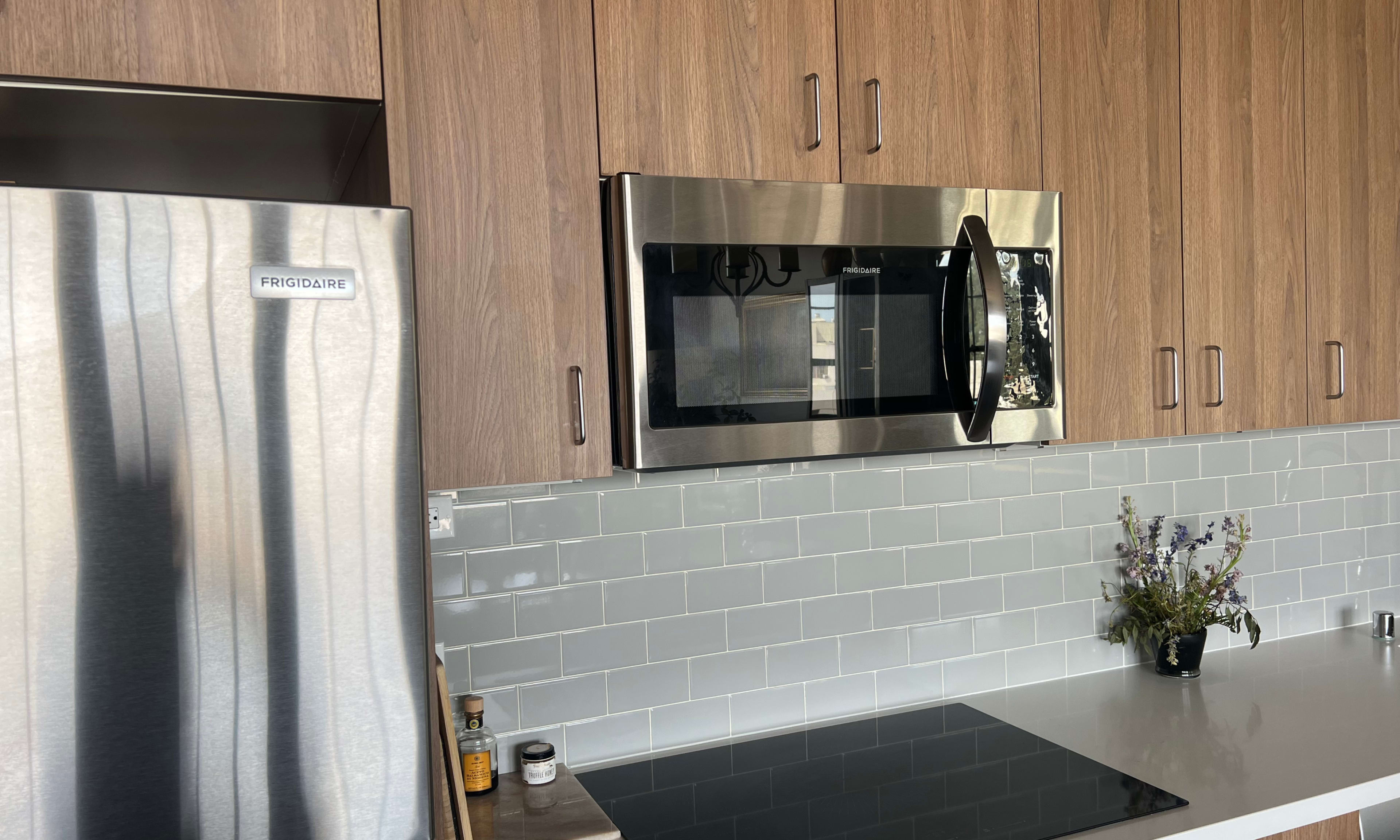 The image shows a modern kitchen with wooden cabinets, a stainless steel microwave above a stove, and a refrigerator labeled "FRIGIDAIRE" next to it.