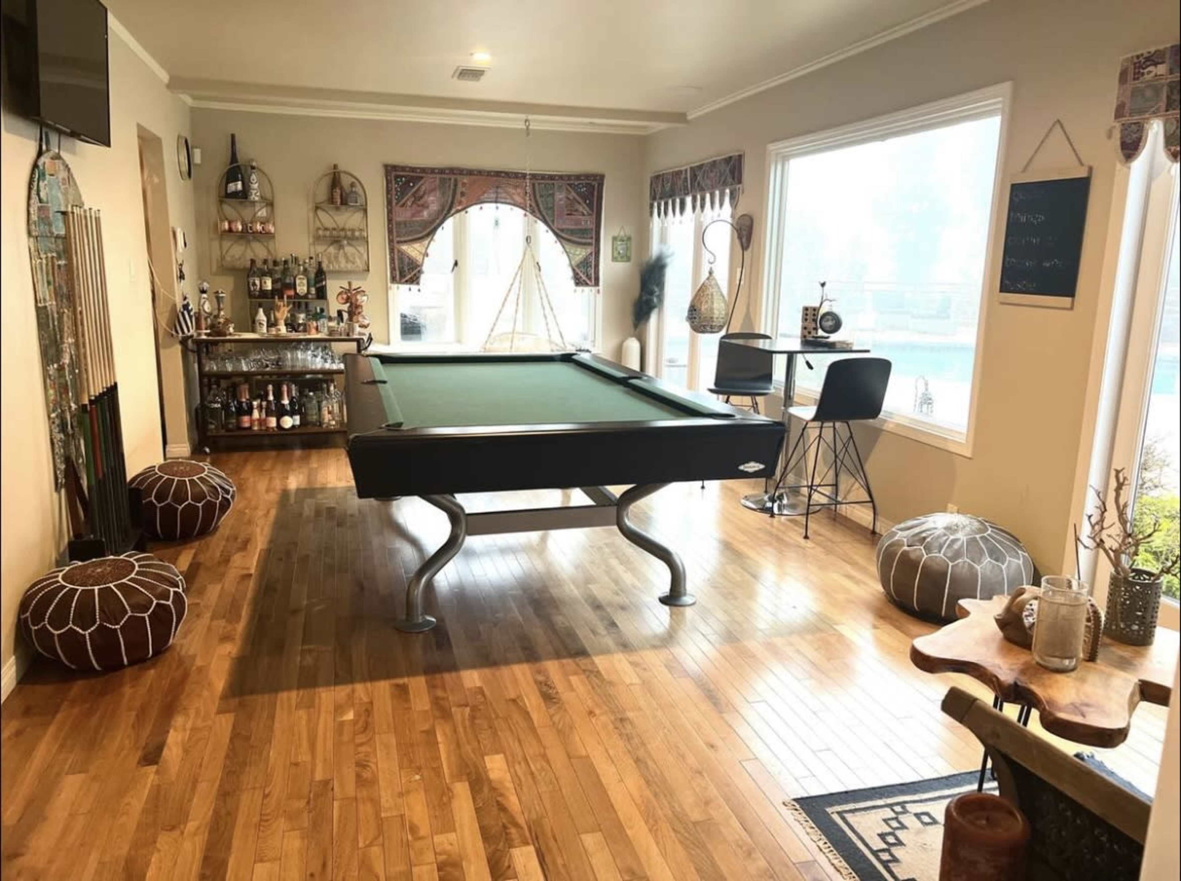 The image shows a spacious game room featuring a pool table in the center, surrounded by decorative elements and a bar area with seating.