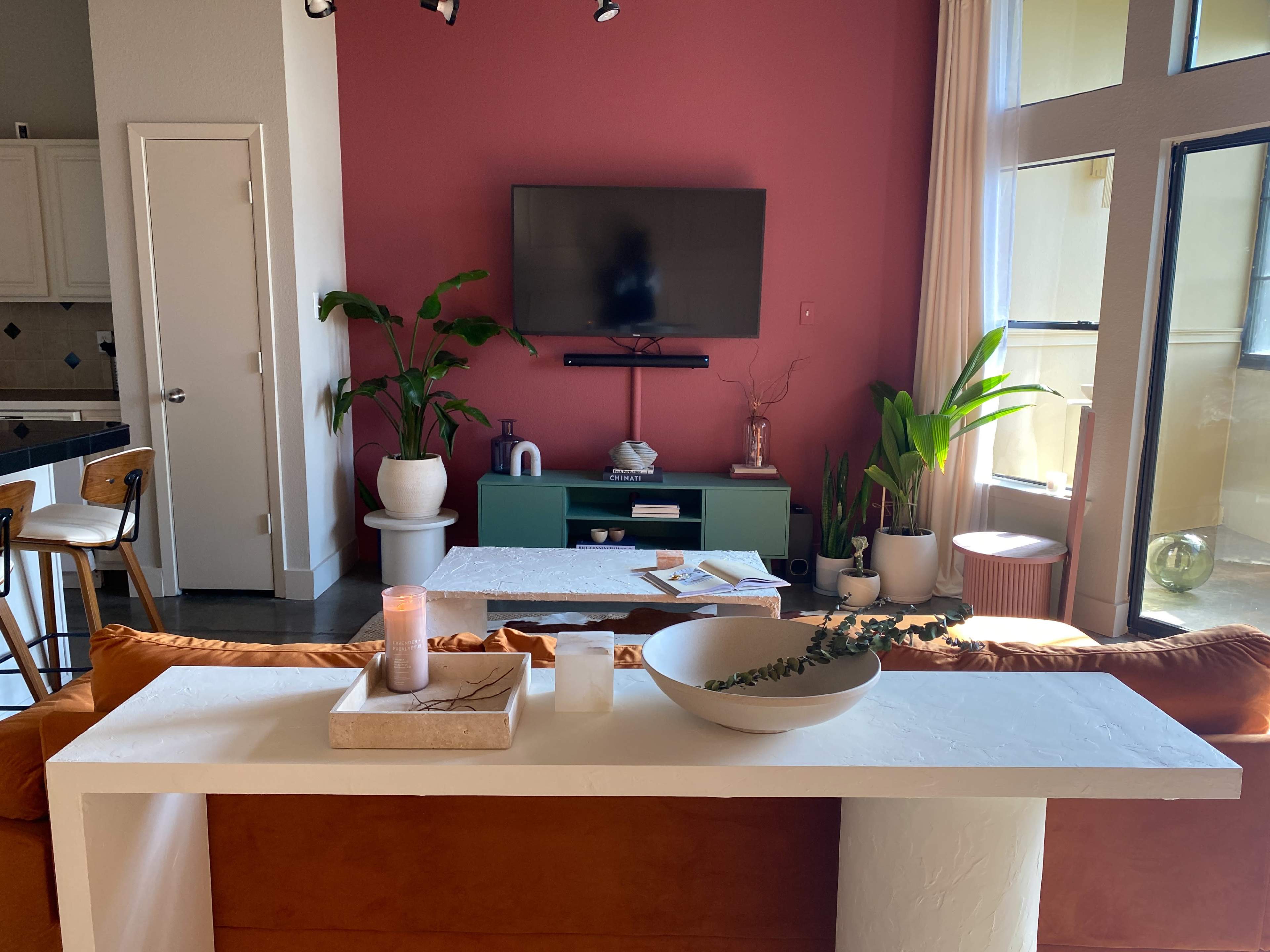 A living room with a television mounted on a wall, surrounded by indoor plants and modern furniture, including a white console table and an orange couch.