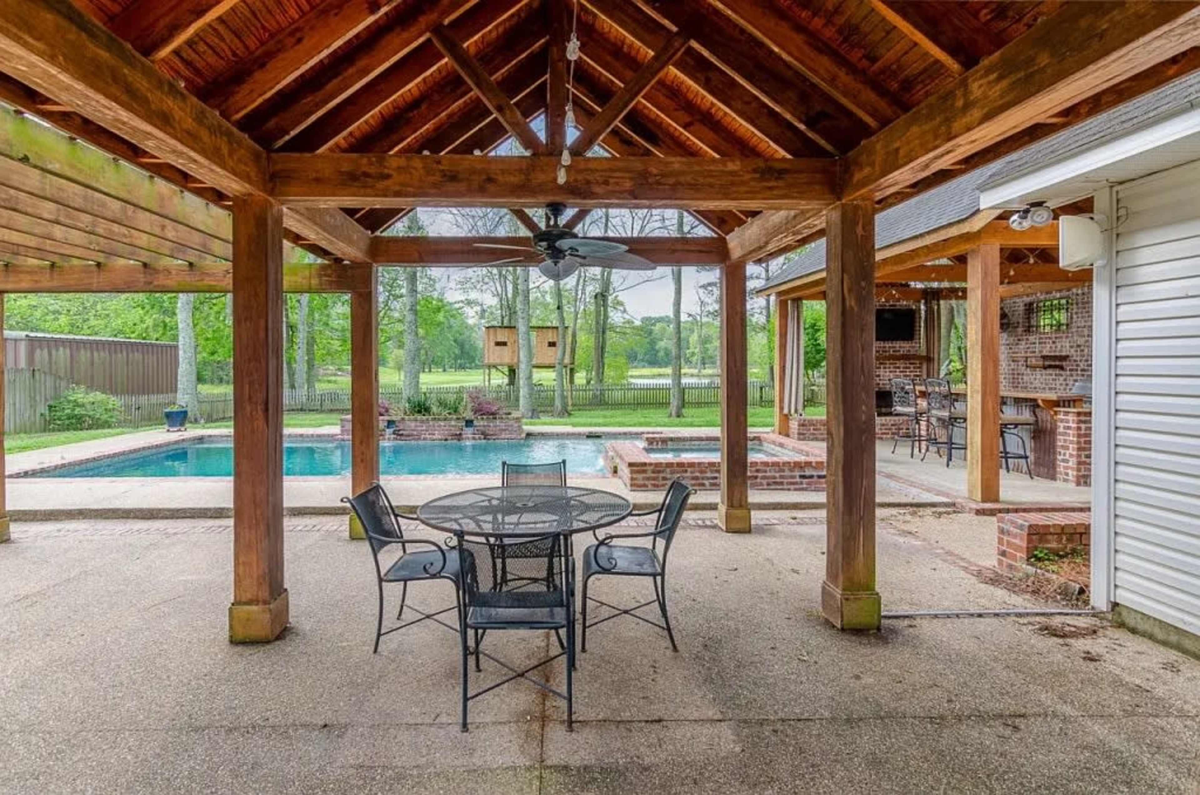 The image shows a covered outdoor patio area with a round dining table and chairs, overlooking a swimming pool and green landscape beyond.