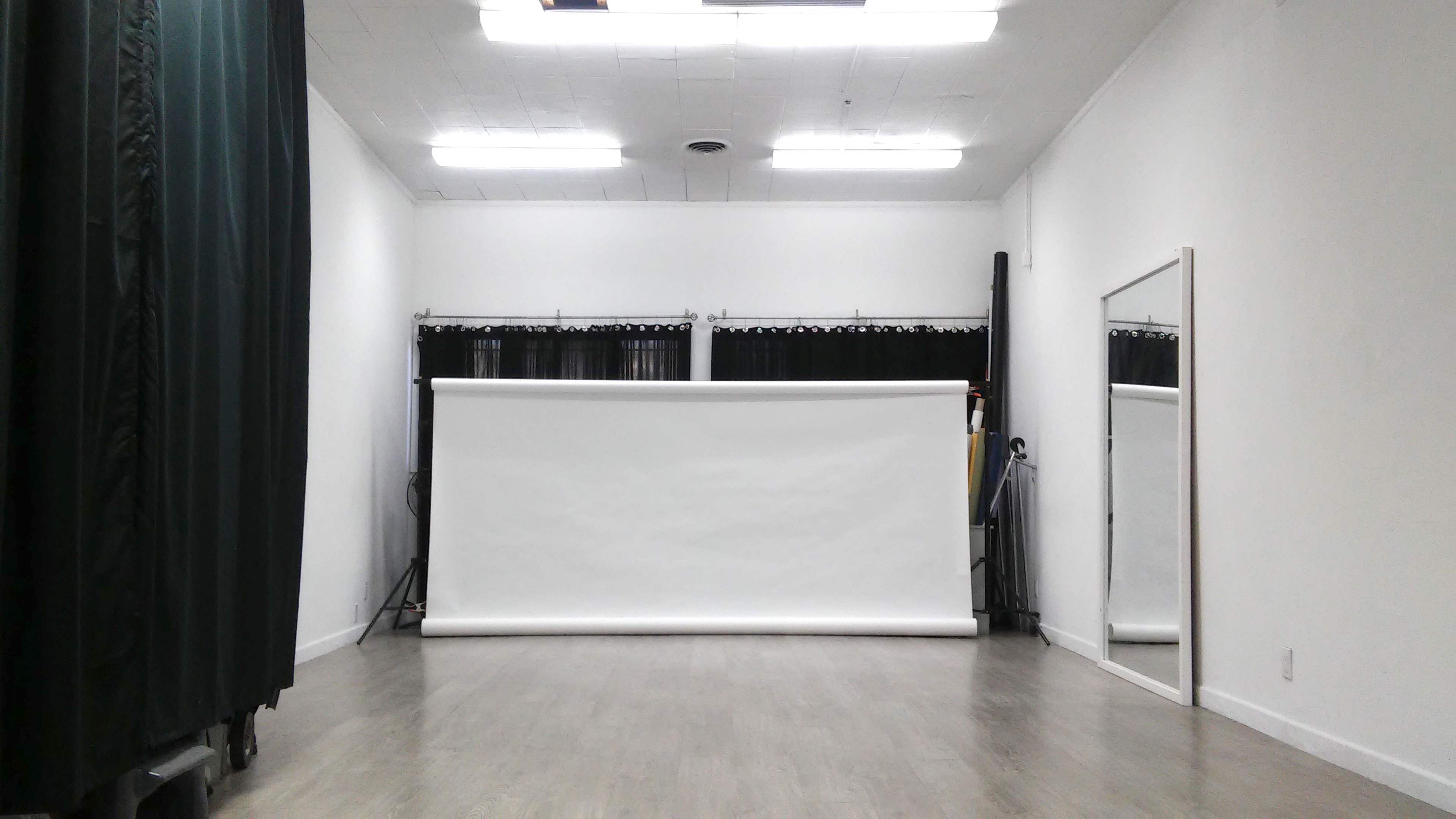A minimalist studio space features a white backdrop on stands, surrounded by dark curtains and a tall mirror against the wall.