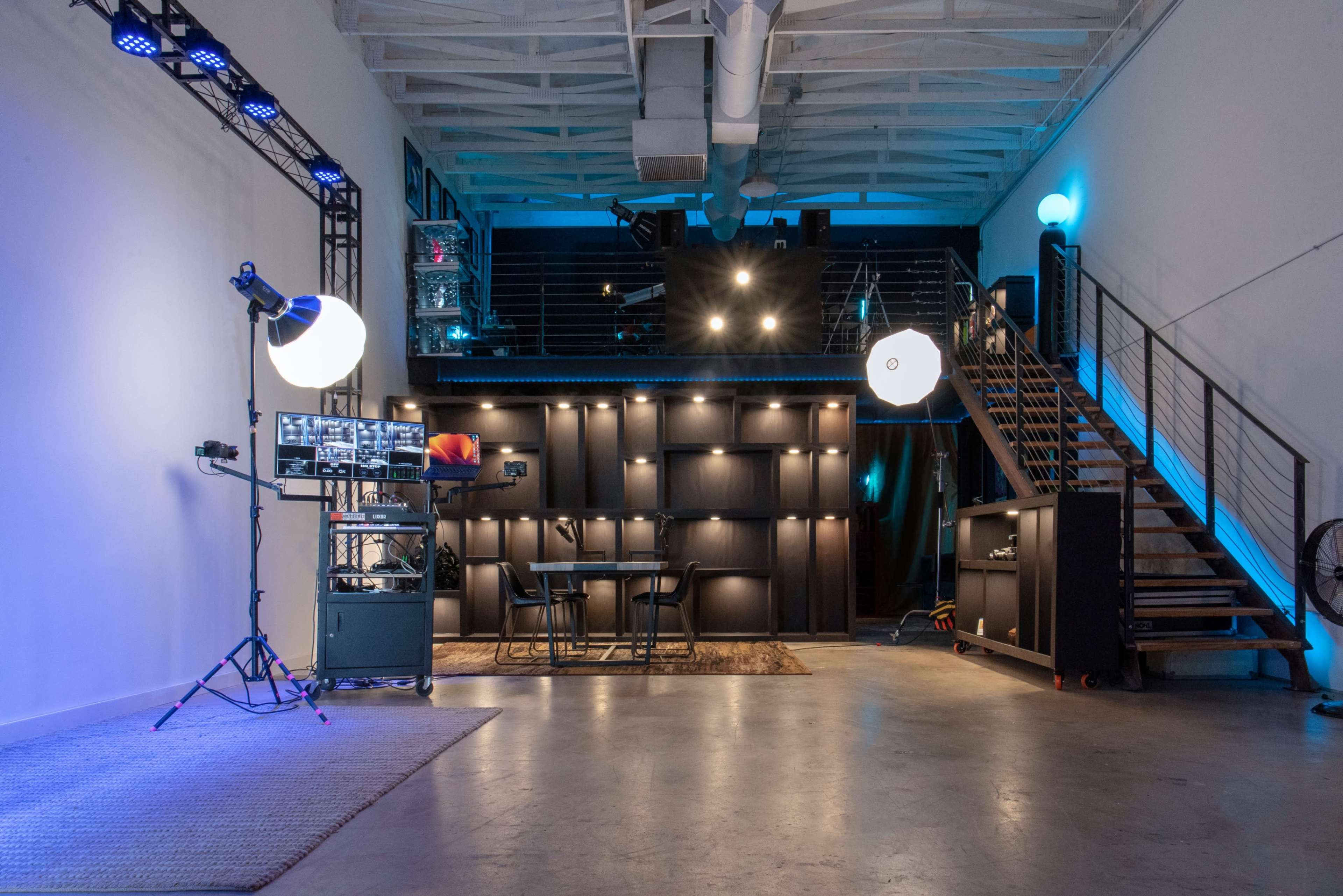 The image shows a spacious interior of a studio featuring modern lighting, a staircase, and a table set against a backdrop of dark cabinetry.