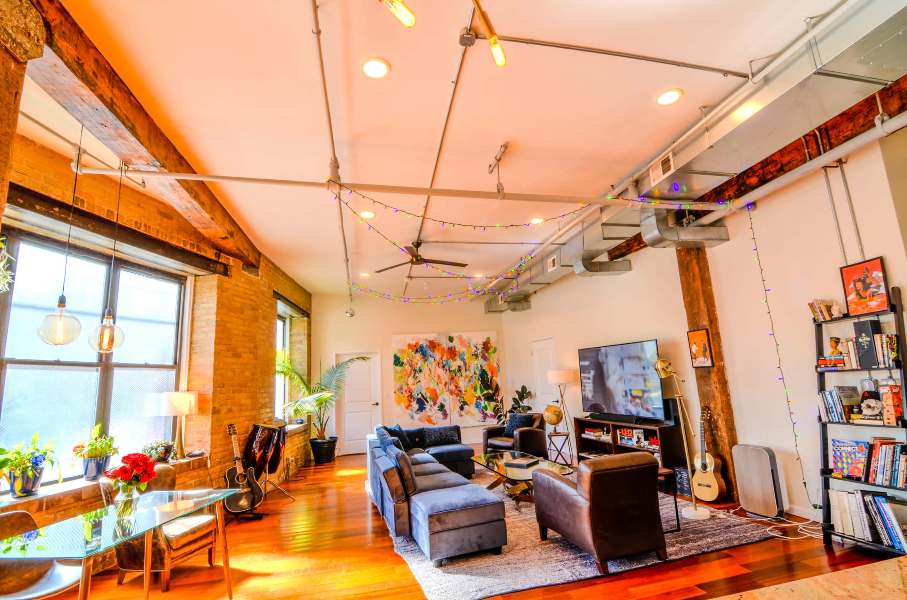 A spacious living room features exposed wooden beams, colorful artwork, a large television, and furniture arranged around a central rug.