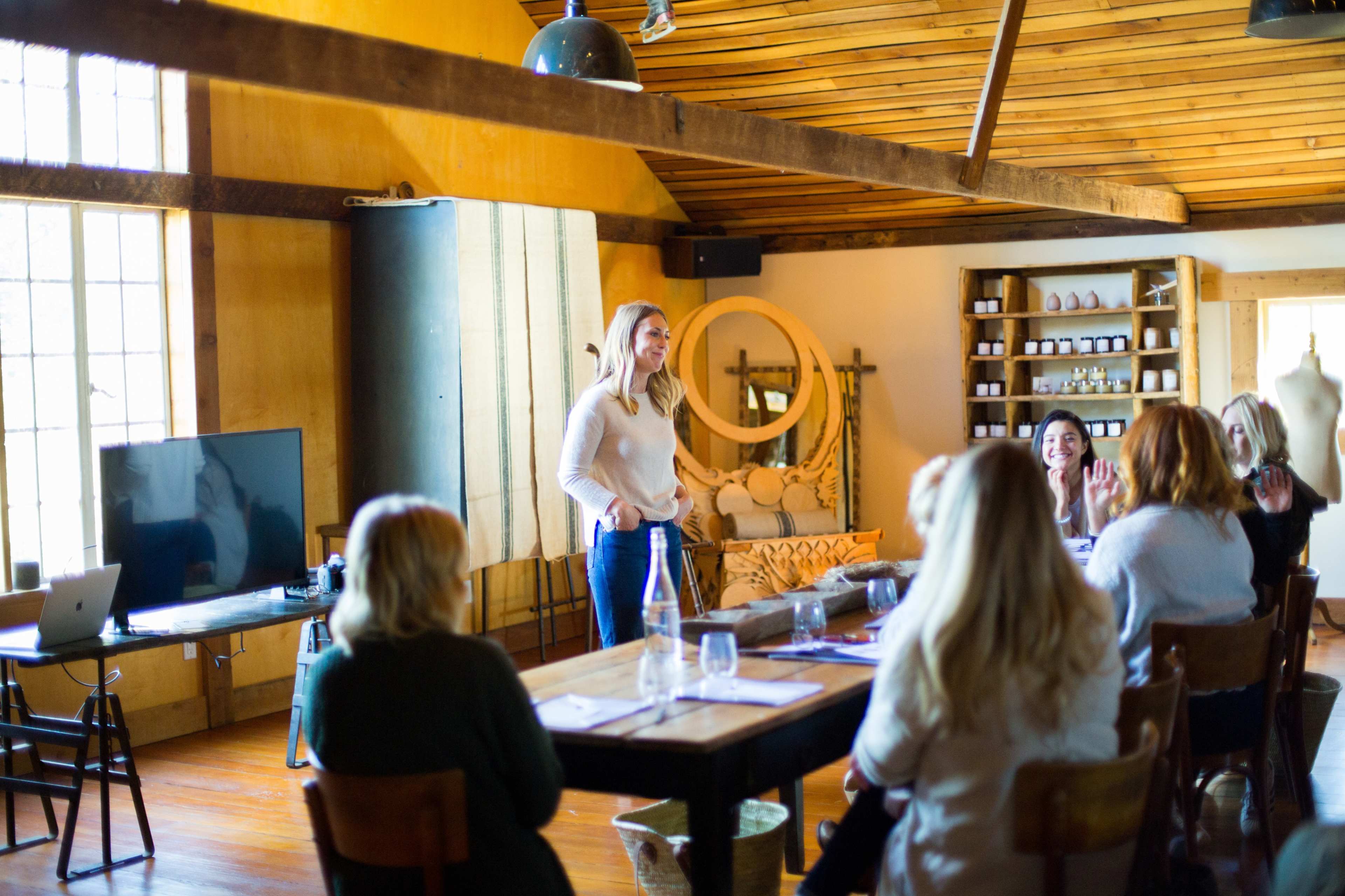 A woman stands in front of a group of seated individuals in a bright, rustic meeting space with wooden beams and shelves.