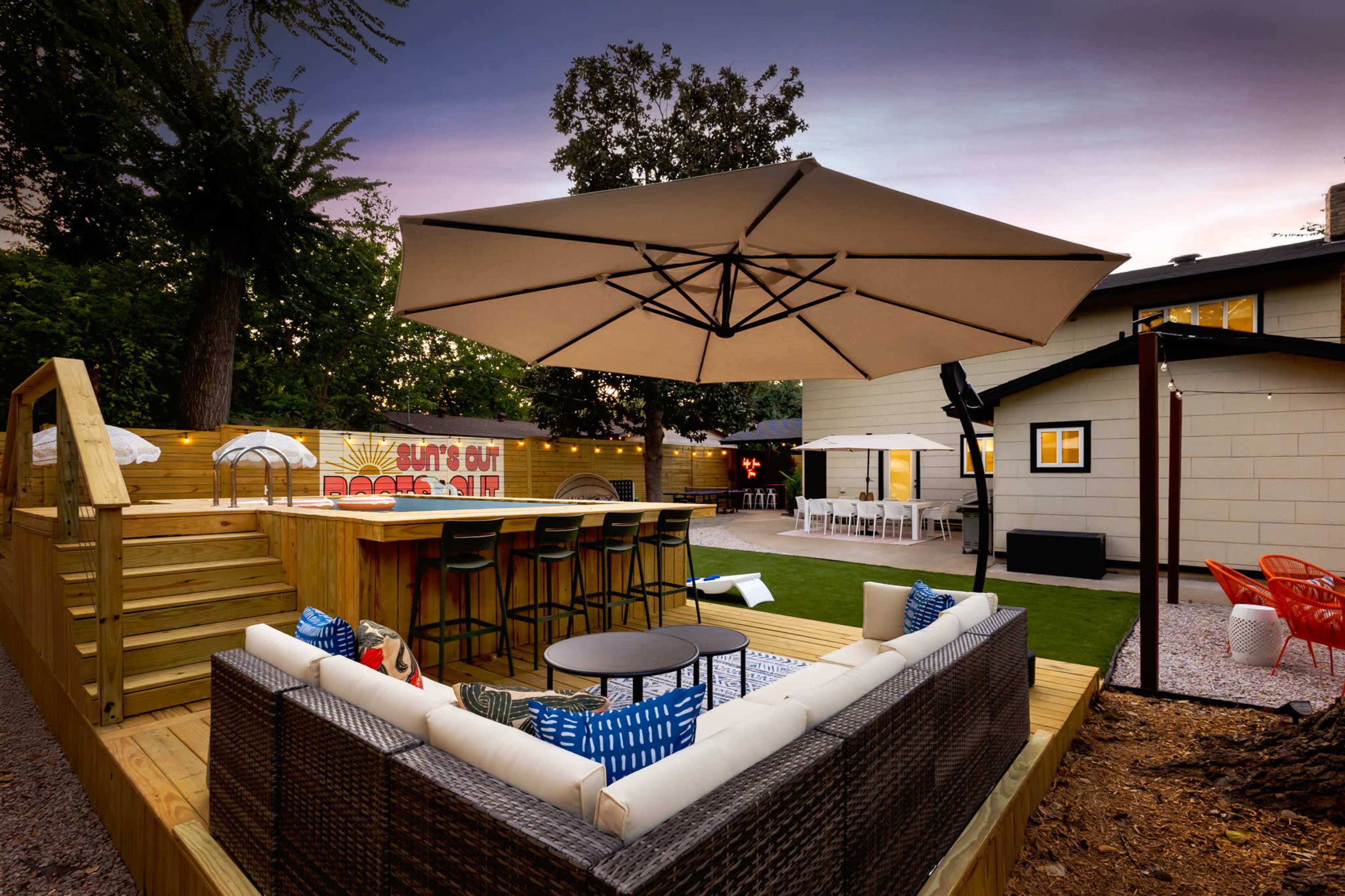The image shows an outdoor patio area with a large umbrella over a seating arrangement and a bar, set against a backdrop of a modern house and landscaped yard.