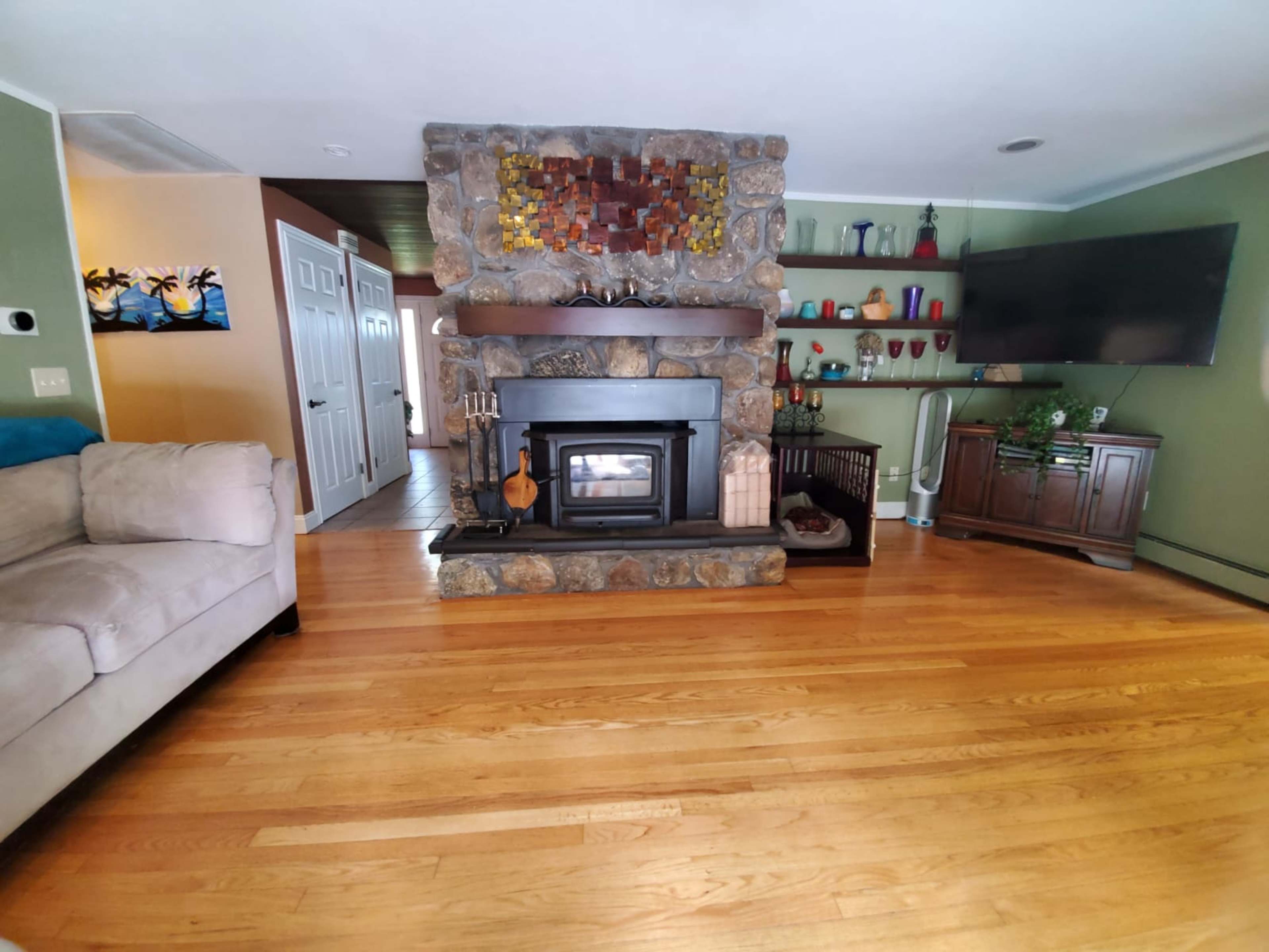 A living room with a stone fireplace, wooden flooring, a television on the wall, and shelves displaying various decorative items.