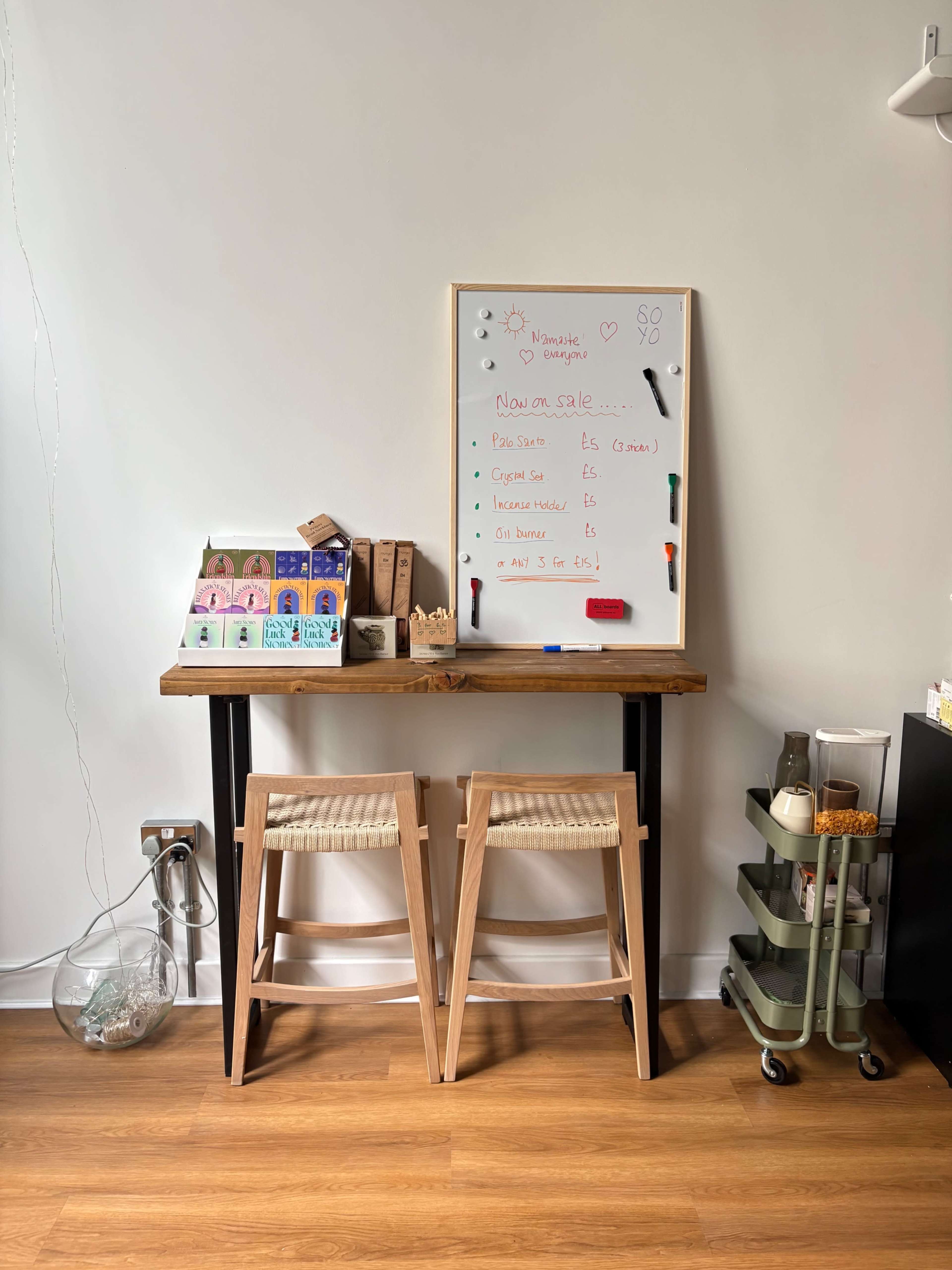 A small wooden table is set against a white wall, featuring a whiteboard with notes and several items organized on the surface and below it.