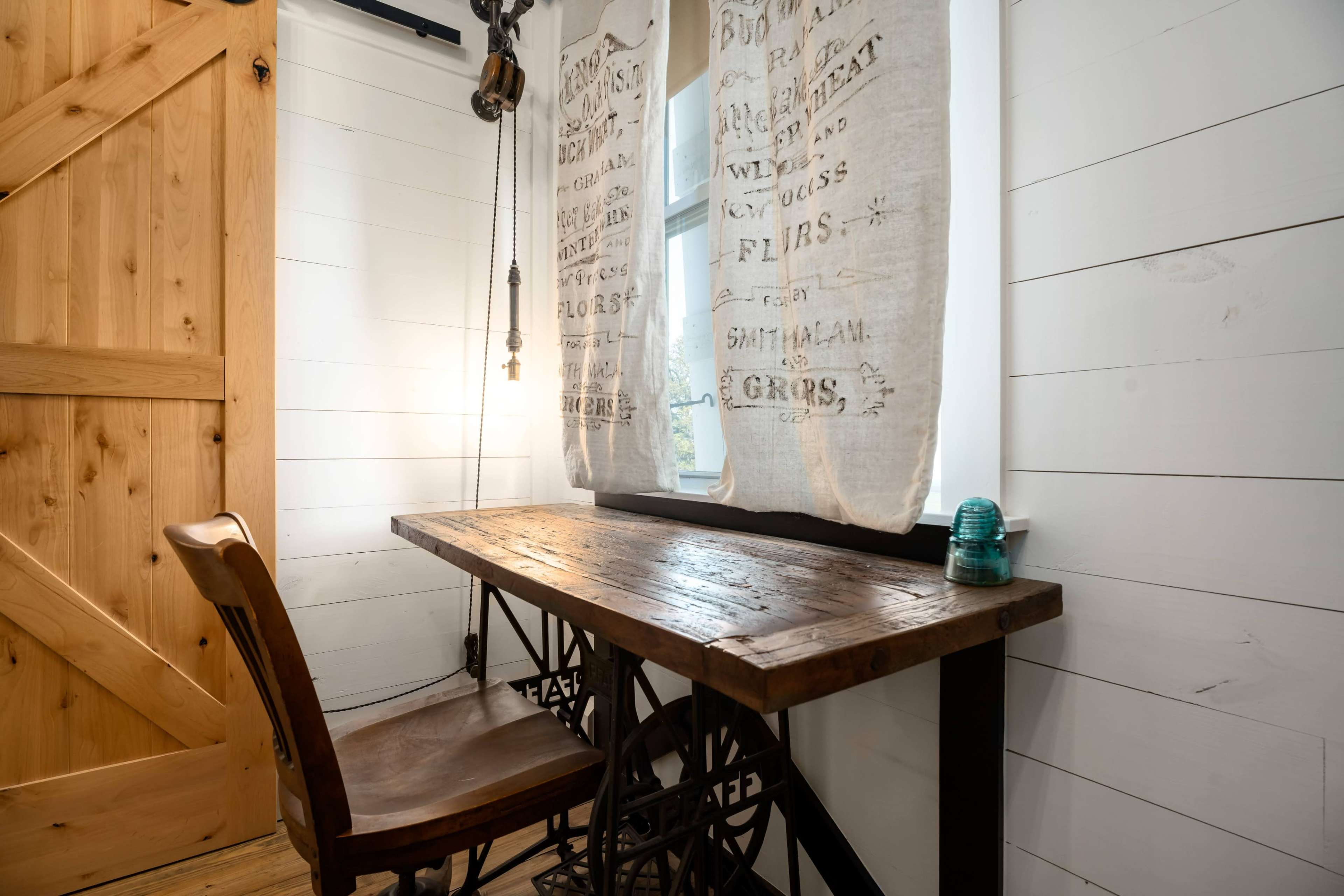 A rustic wooden desk with a vintage chair is positioned against a wall adorned with light-colored wooden panels and sheer curtains.