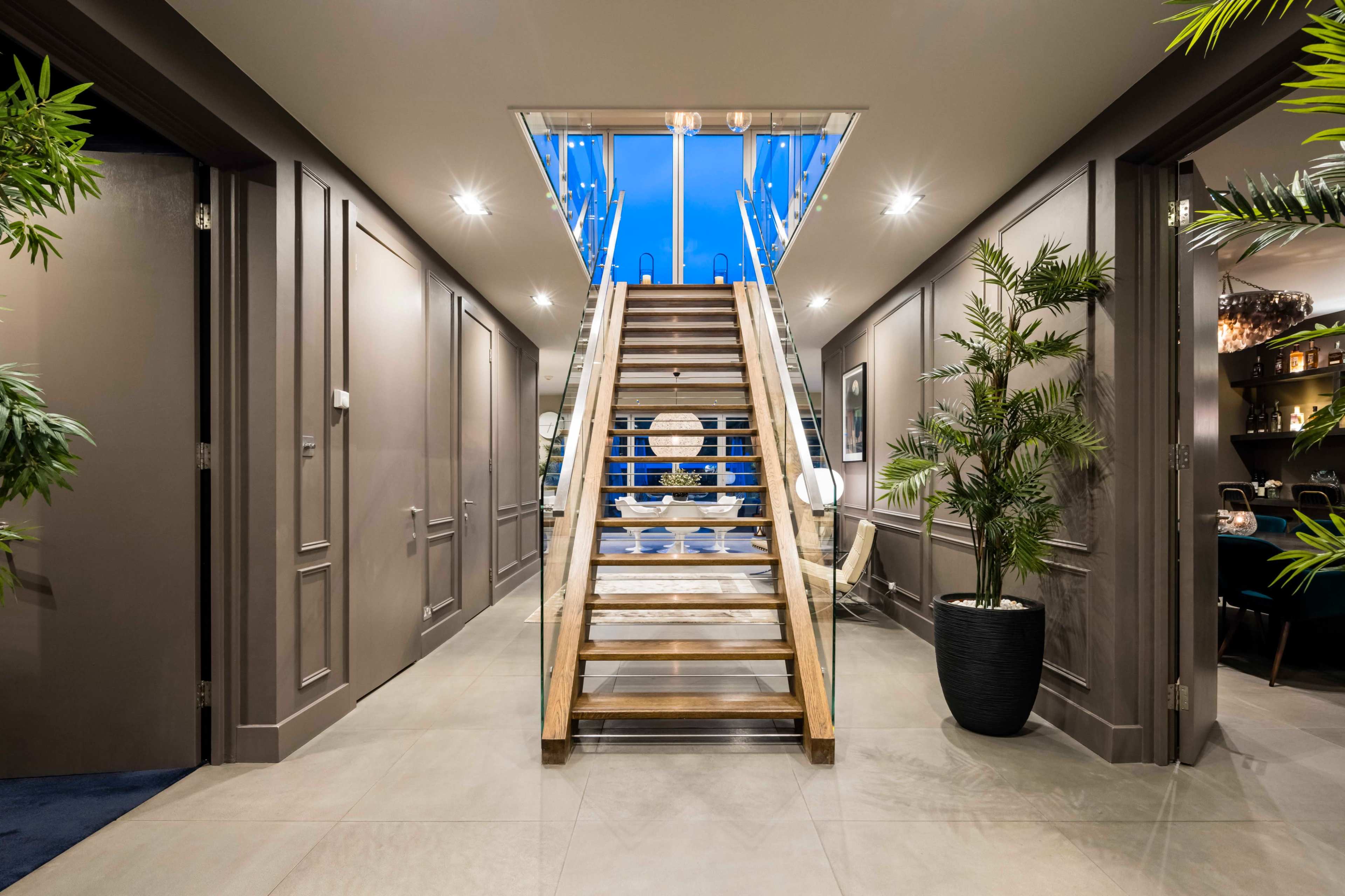 A wooden staircase with a glass rail ascends from a well-lit hallway featuring green plants and stylish decor.