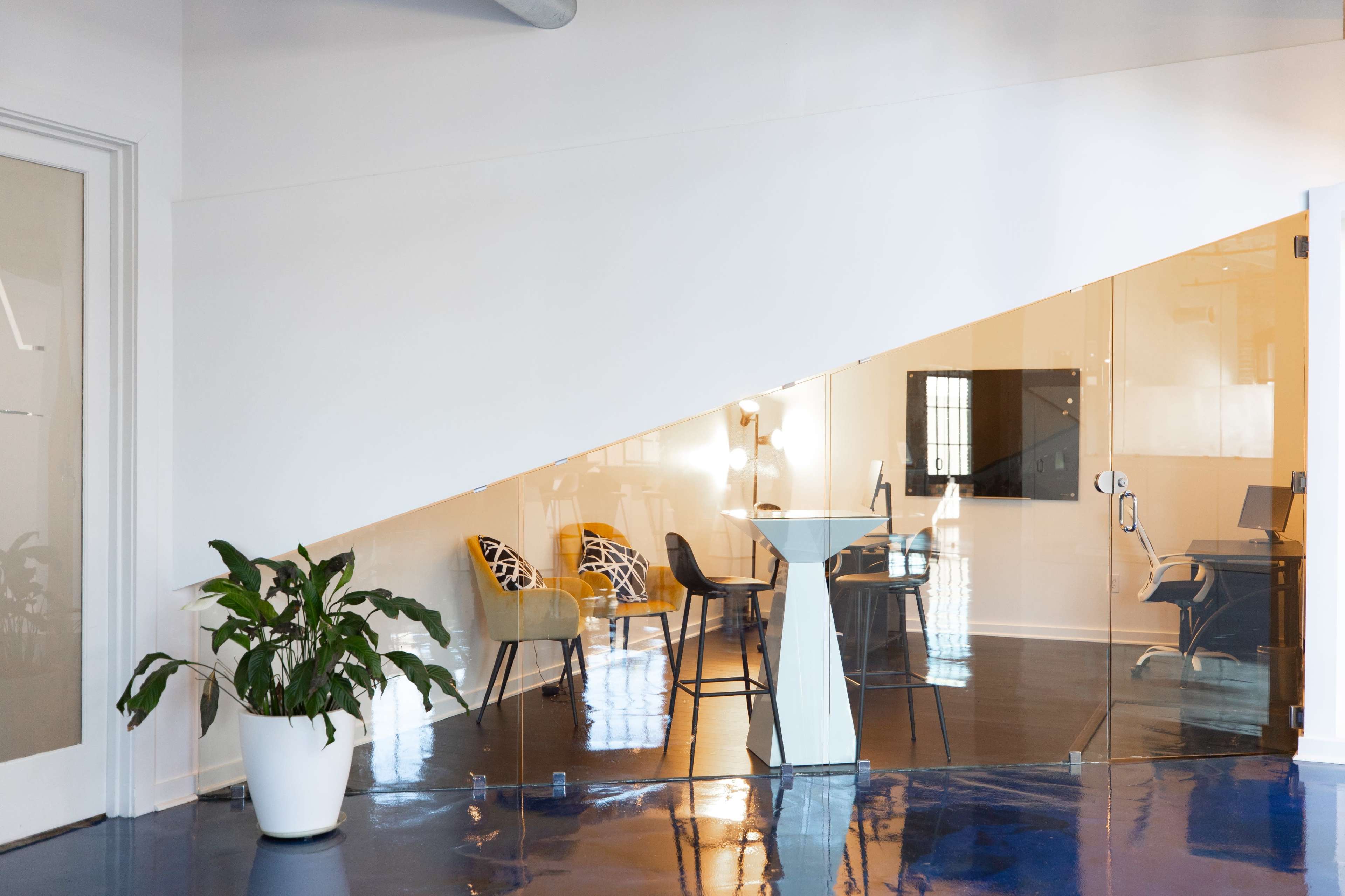 A modern office space features a glass-walled meeting area beneath a staircase, with seating arrangements and a potted plant in the foreground.