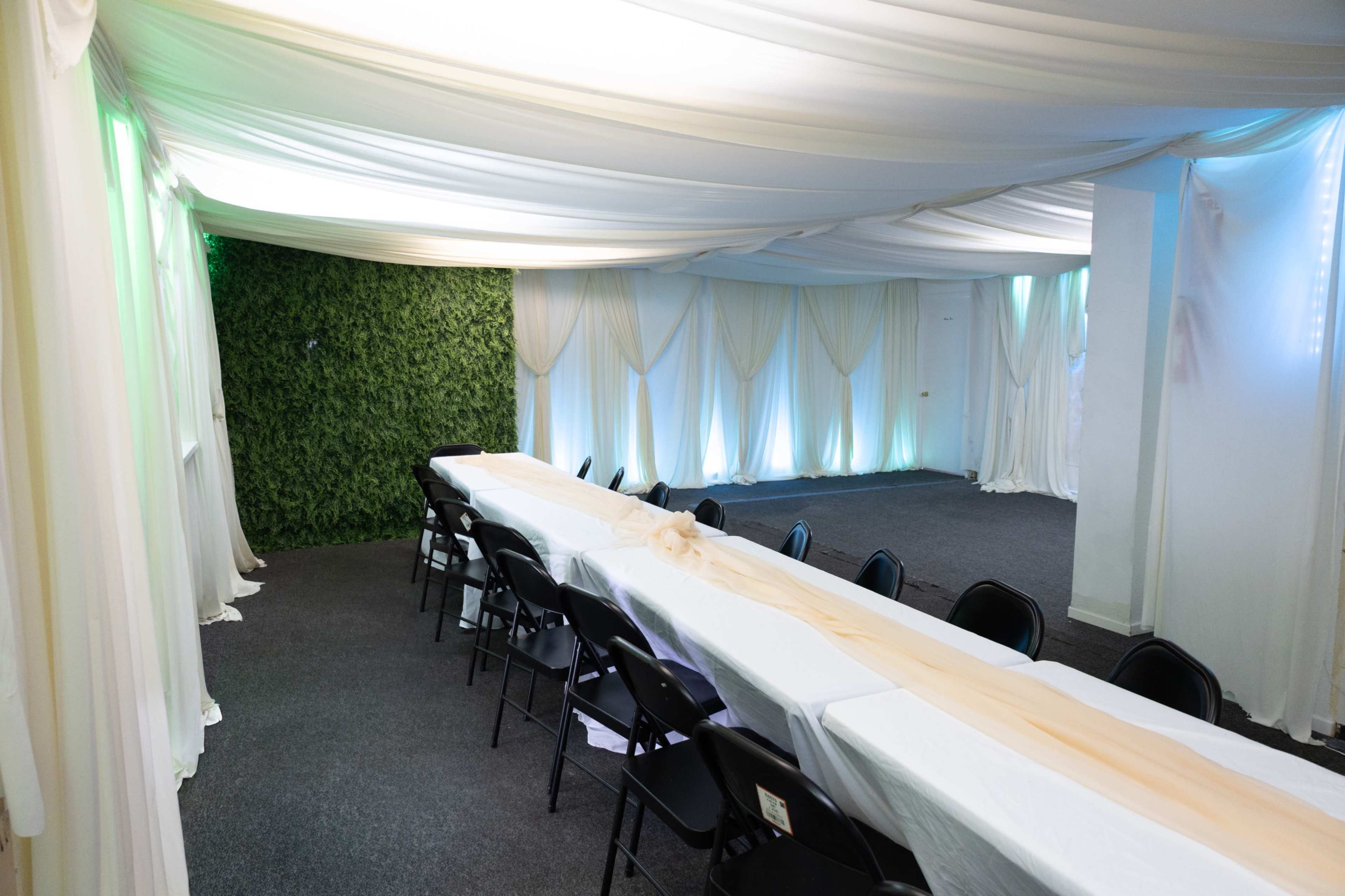 The image shows a banquet-style room with a long table covered in white tablecloths, surrounded by black chairs, and draped with white fabric and green foliage accents.