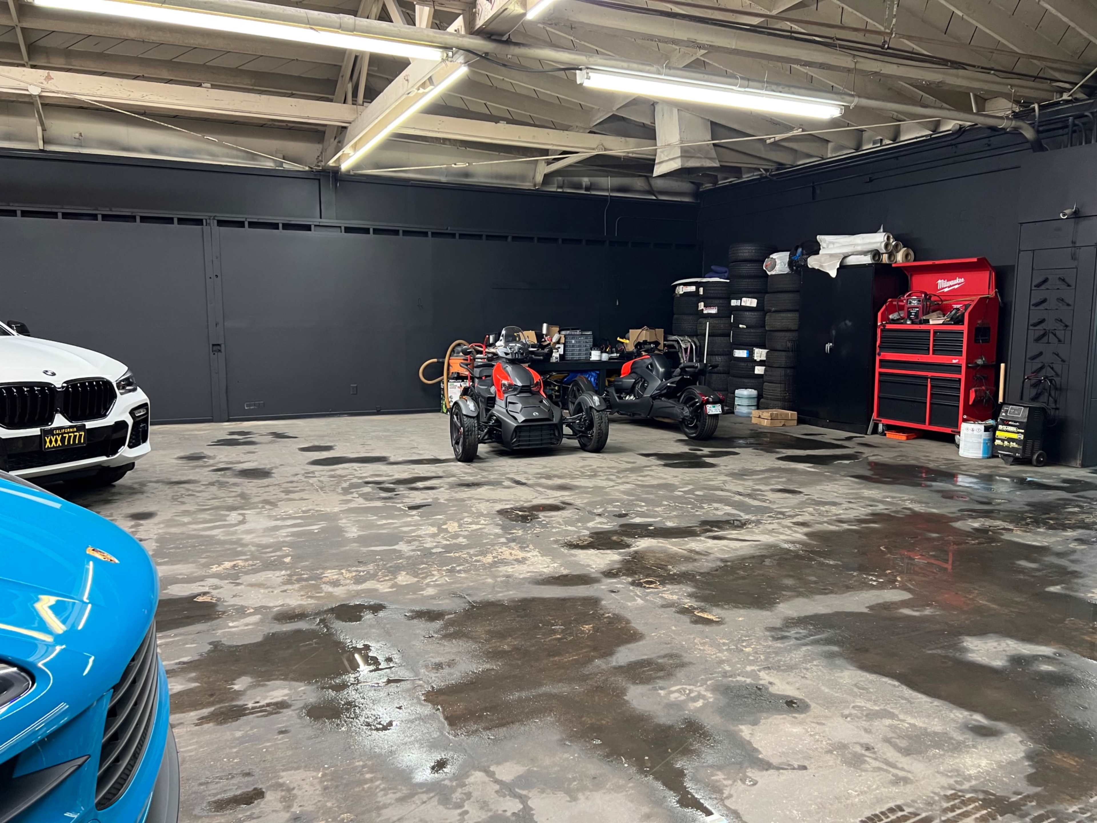 An empty garage space with a blue car on the left, a black and red motorcycle in the center, tool boxes, and stacks of tires against a black wall.