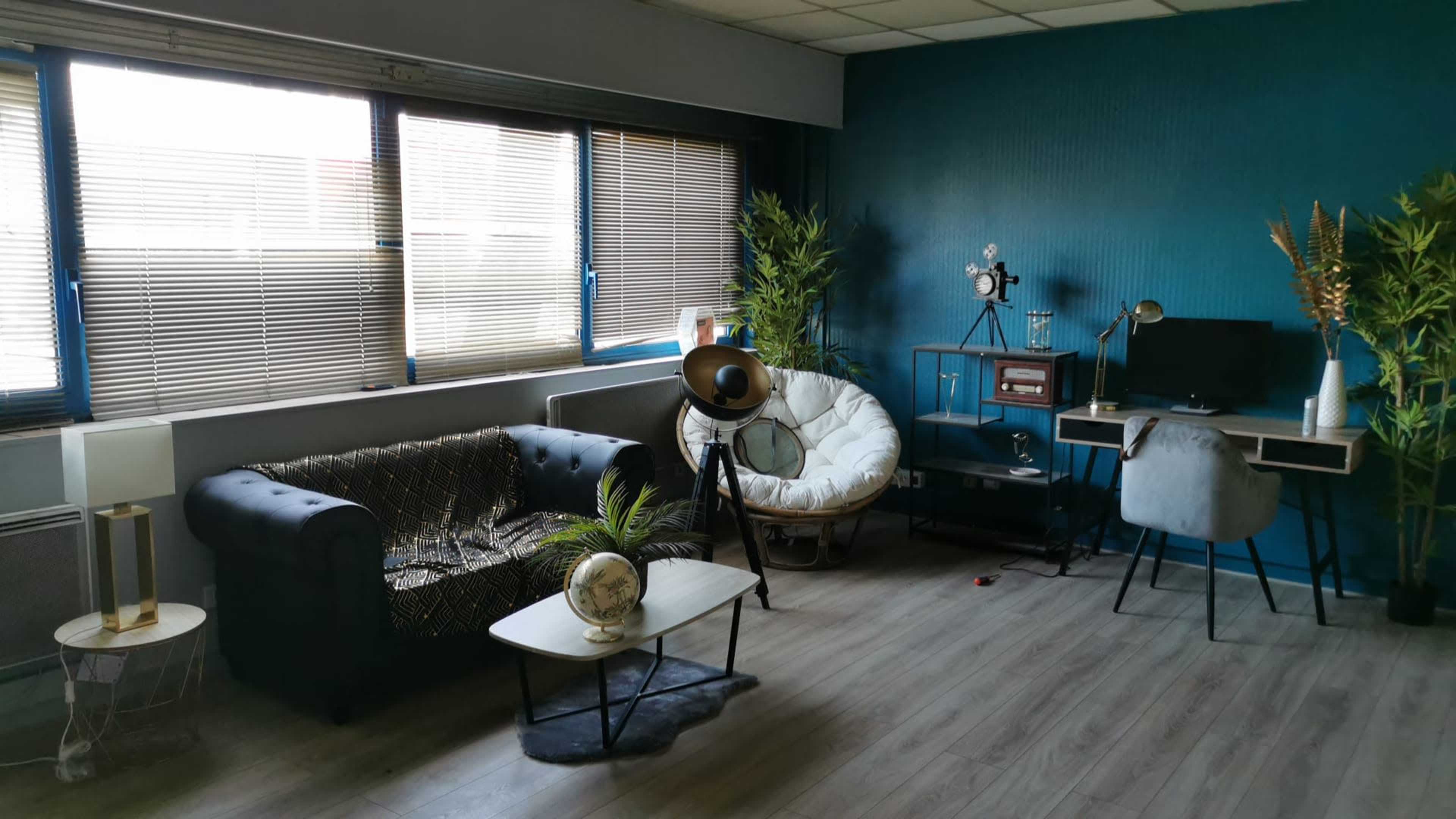 A modern, stylish room featuring a dark sofa, a round armchair, a small coffee table, a desk with a computer, and decorative plants against a teal wall.