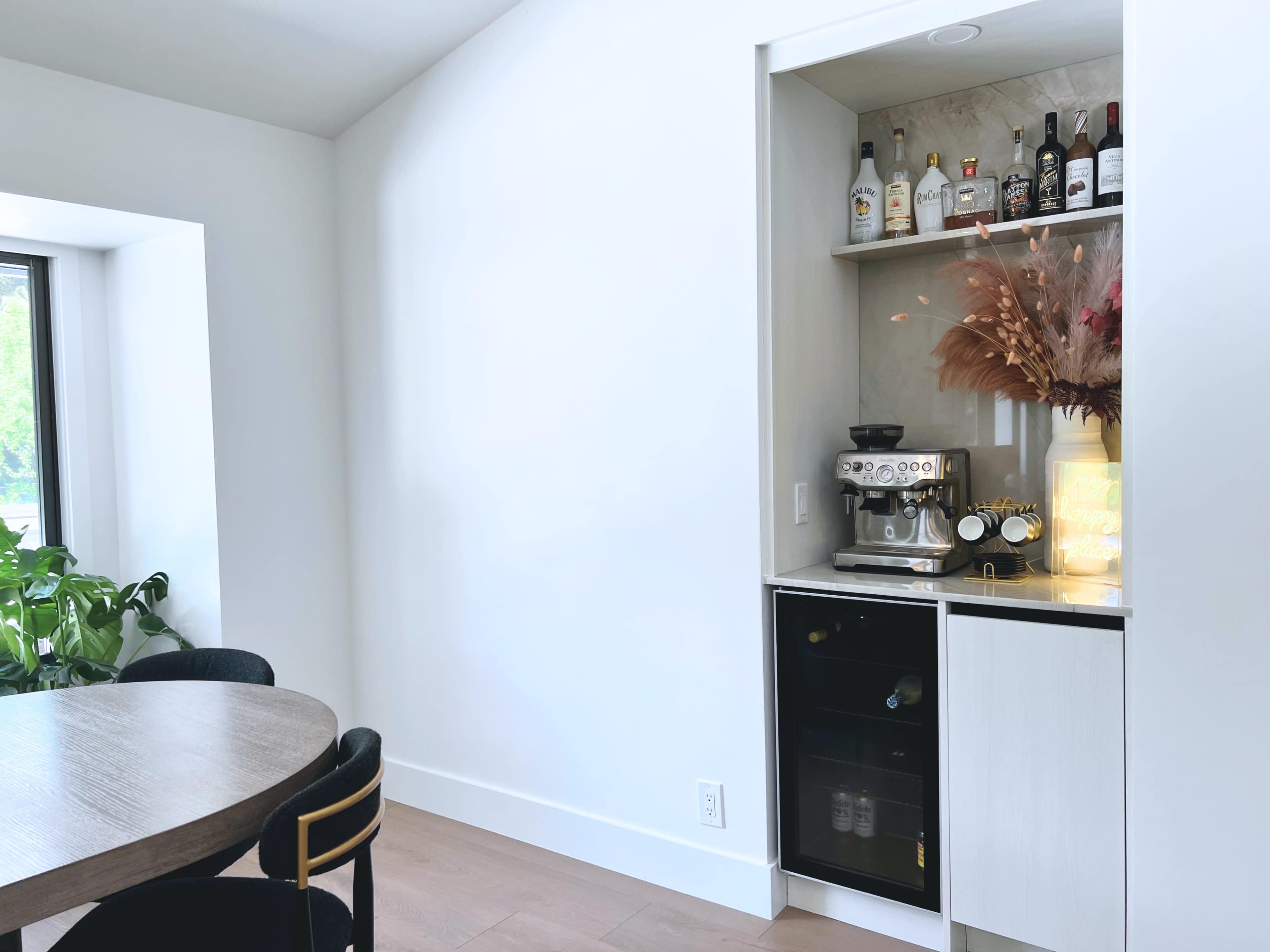 The image shows a modern corner of a room featuring a built-in bar with shelves holding various bottles, an espresso machine on top, and a small refrigerator below, next to a round dining table and a large window.