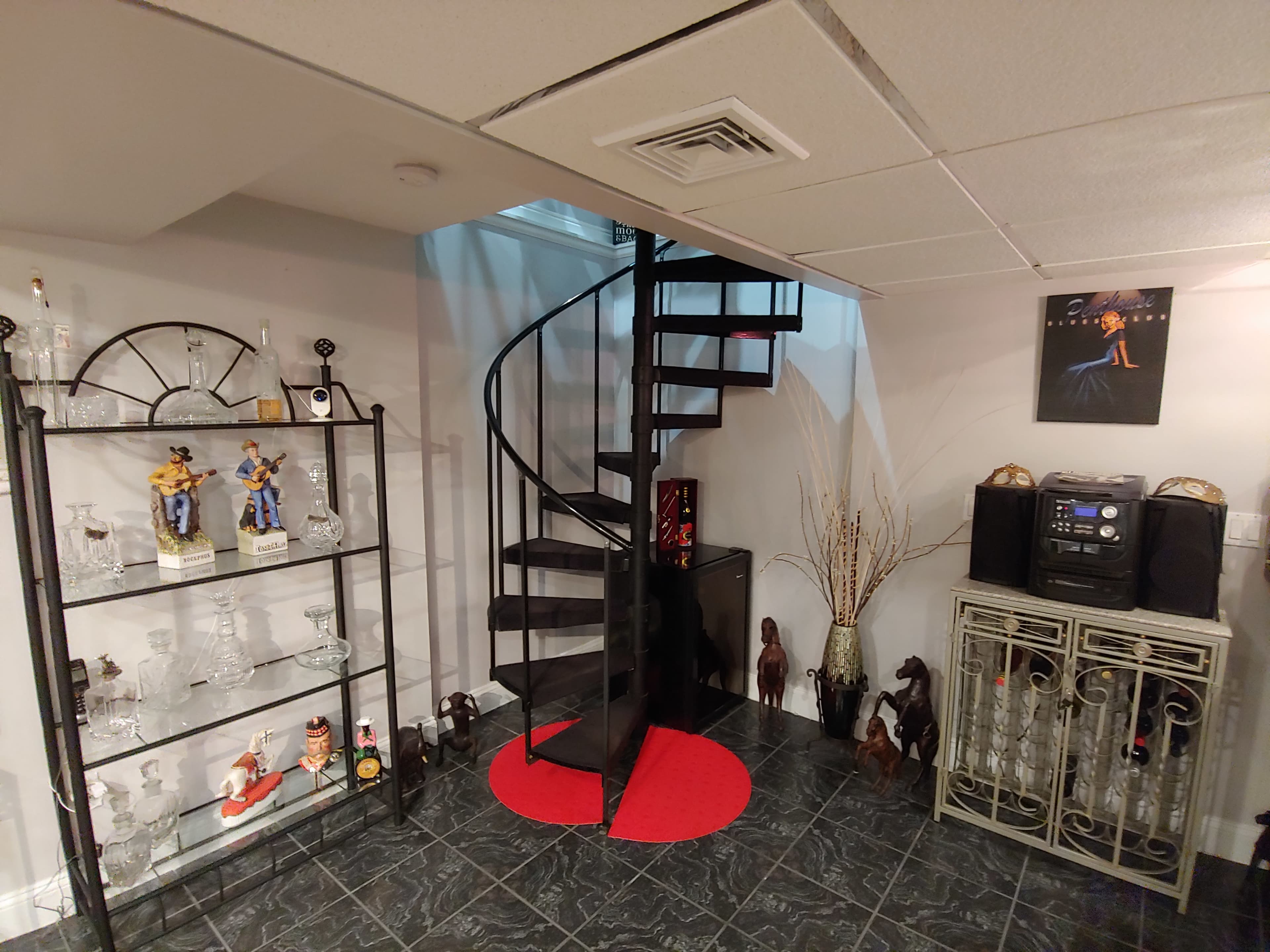 A basement area with a spiral staircase, a round red carpet, decorative glassware on shelves, and various figurines and a stereo system arranged on the floor.