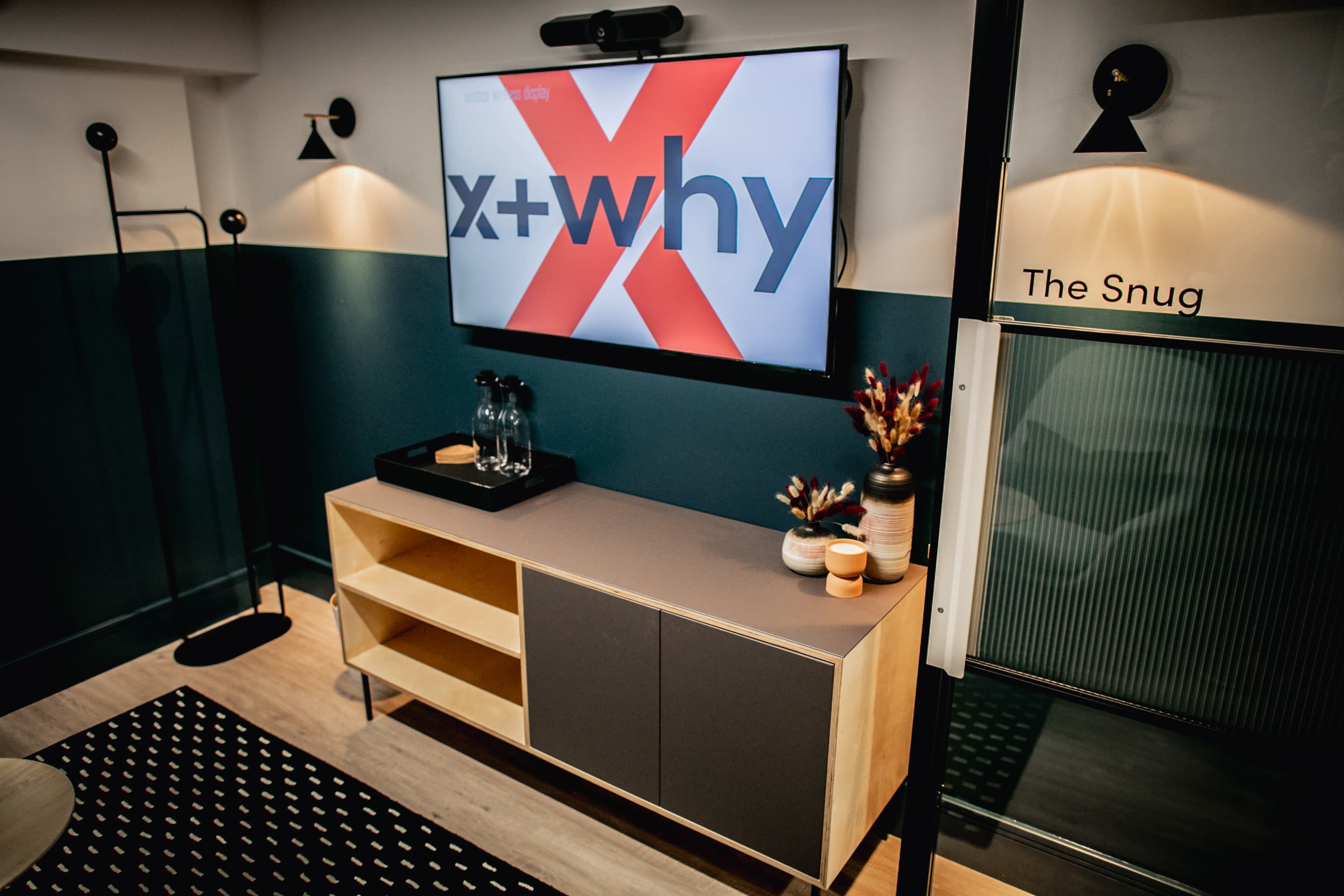 A modern room with a television displaying "x+why," a wooden cabinet holding decorative items, and stylish wall lighting.