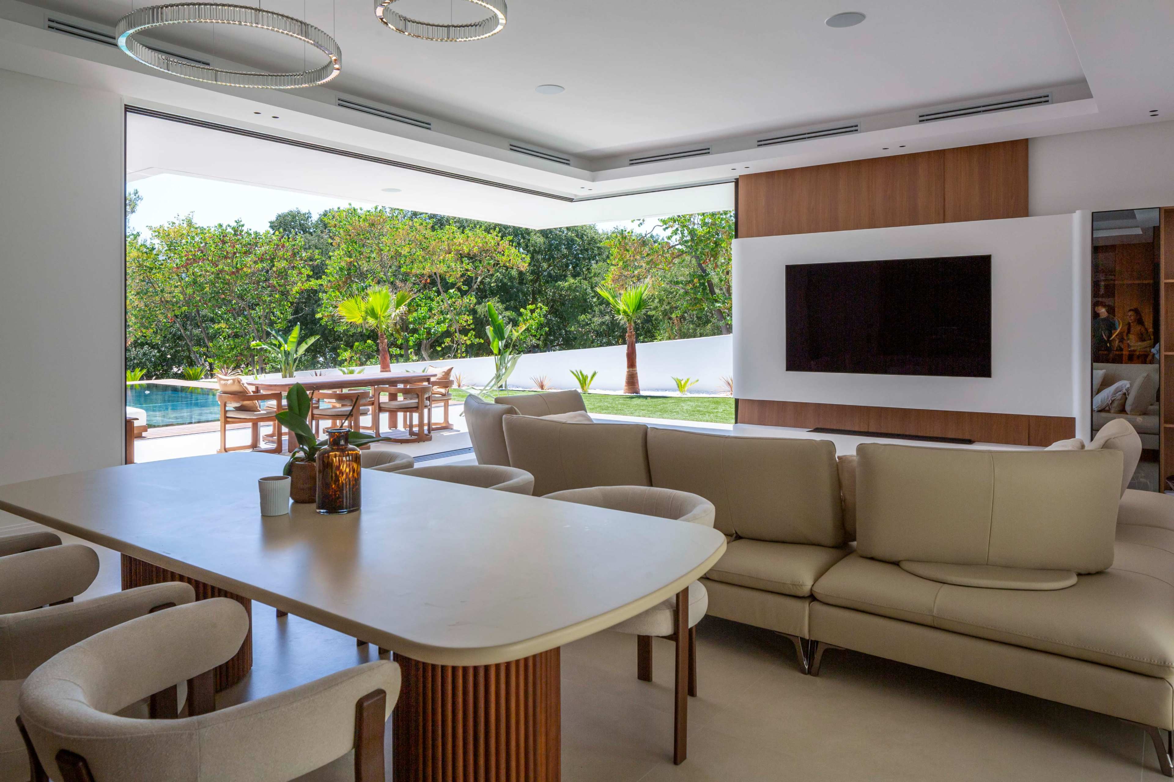 The image shows a modern living room with a white sofa and a dining table, overlooking a pool area surrounded by greenery.