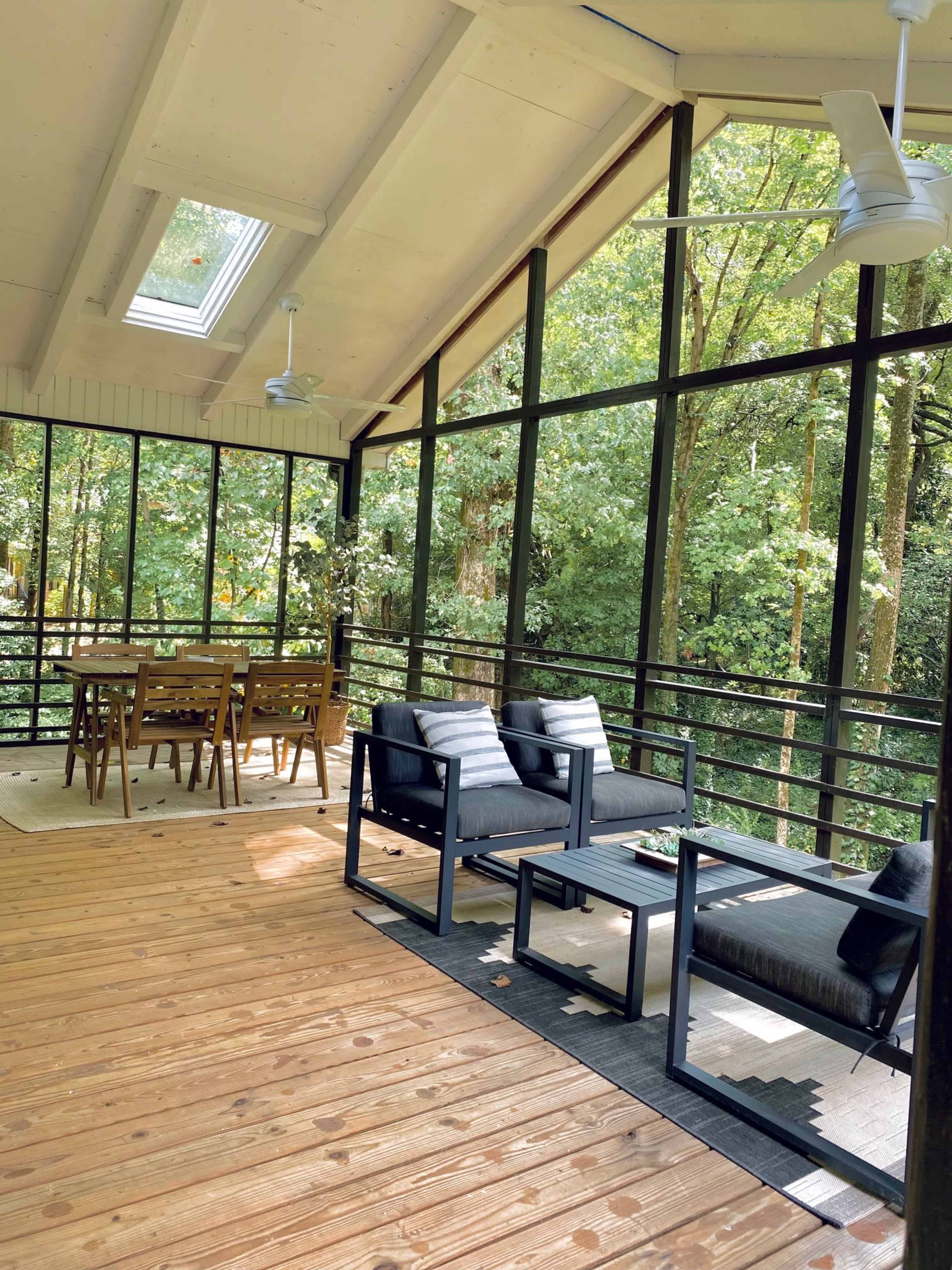 The image shows a screened-in porch with wooden flooring, contemporary furniture, and a dining area overlooking a wooded landscape.