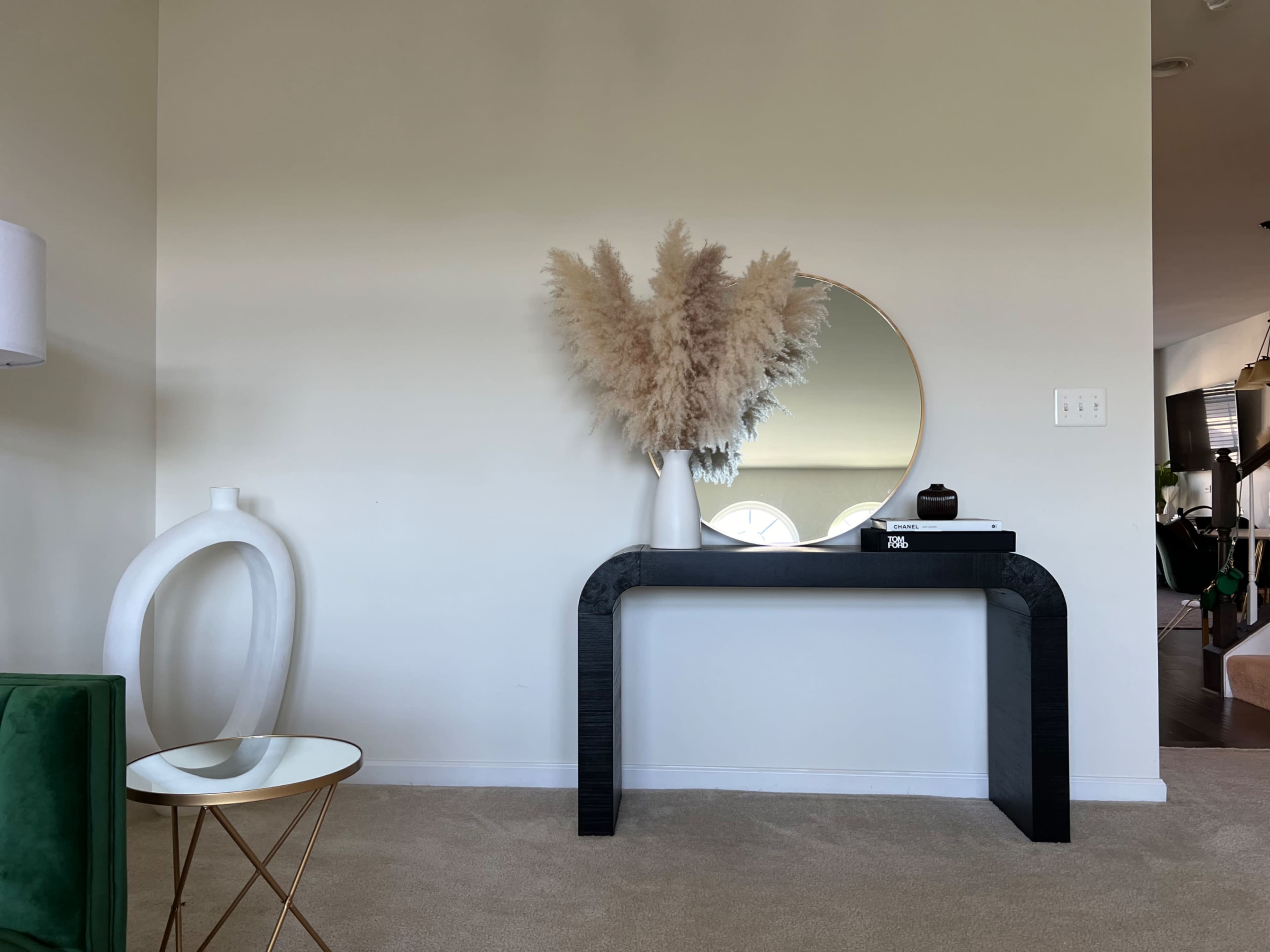 A black console table with a round mirror above it displays a vase of dried pampas grass, accompanied by a decorative white sculpture and a small round table with a glass top.