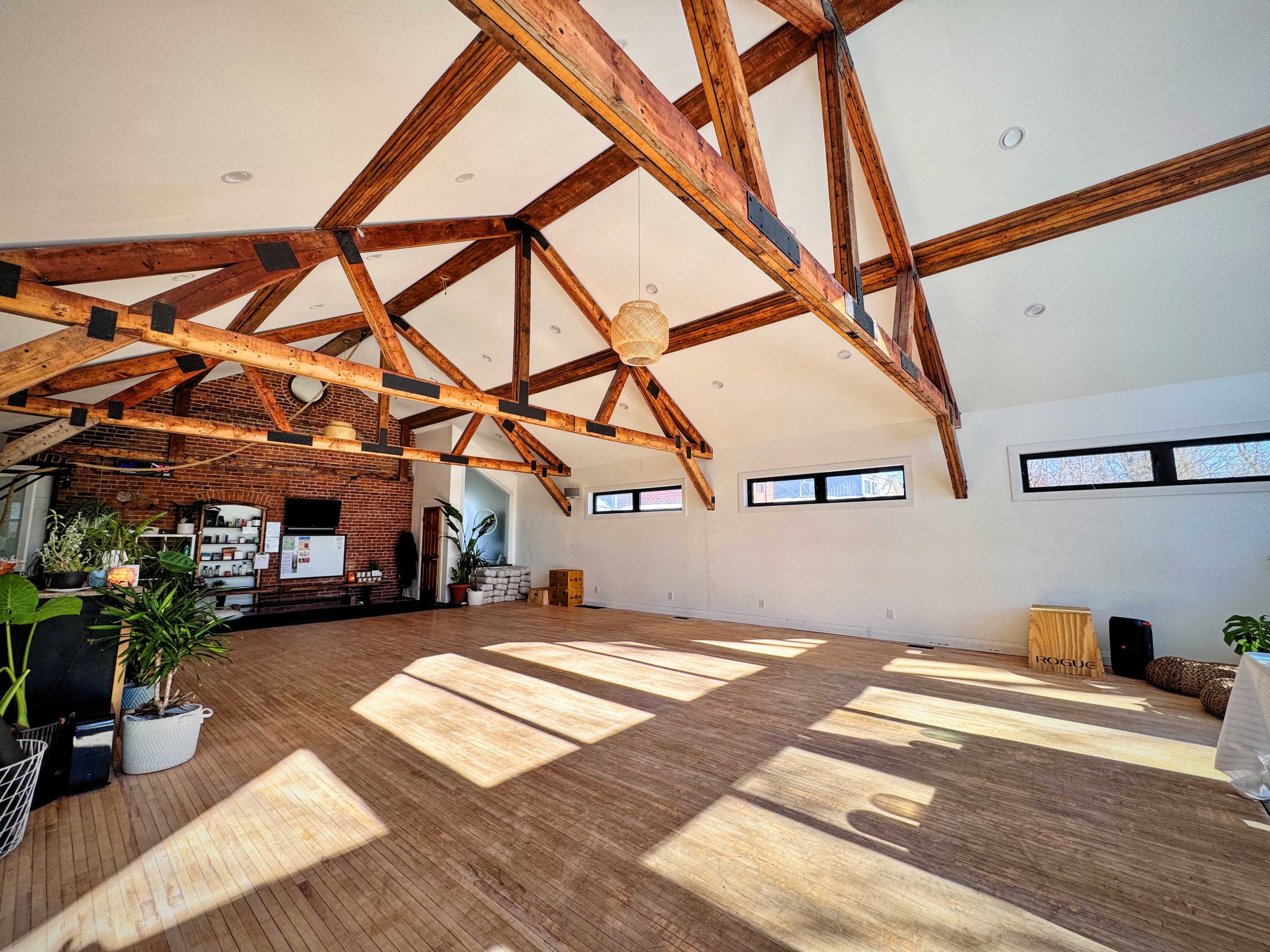The image shows a spacious interior with wooden beams, ample natural light, and hardwood flooring, featuring a minimalist design.