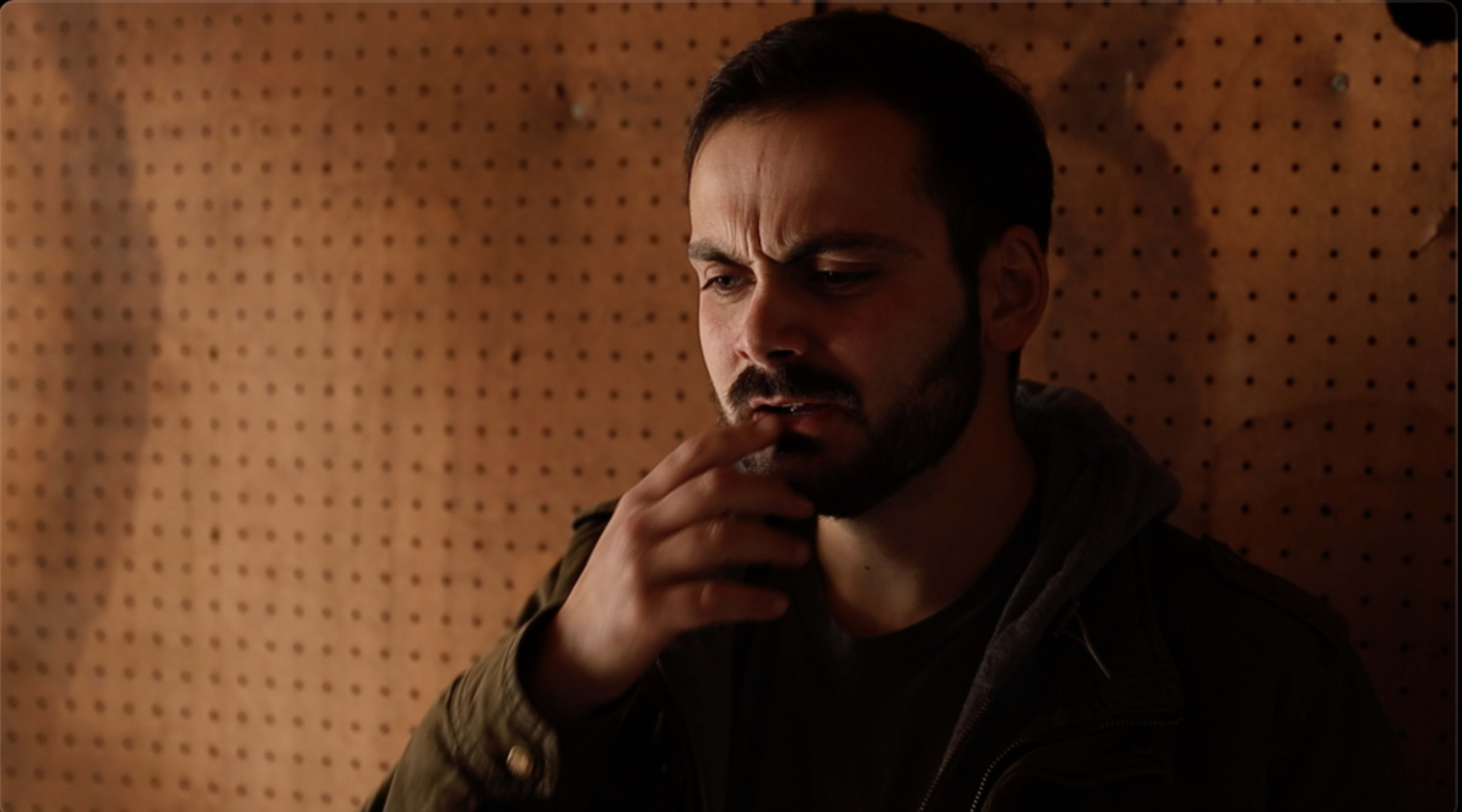 A man with a beard stands in front of a pegboard, touching his chin with a contemplative expression.