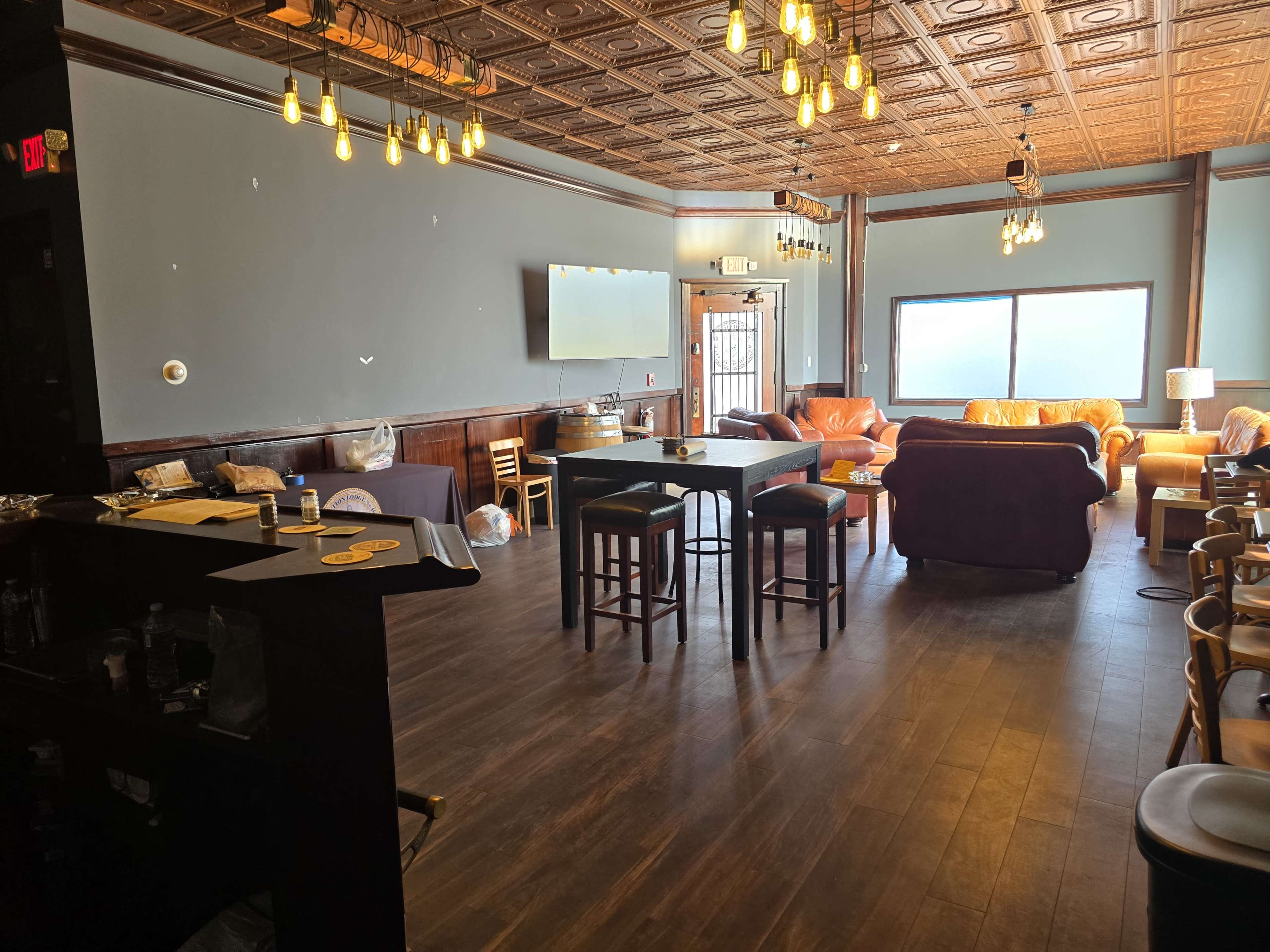 The image shows a spacious lounge area with wooden flooring, a bar on the left, several tables and chairs, and a large window providing natural light.