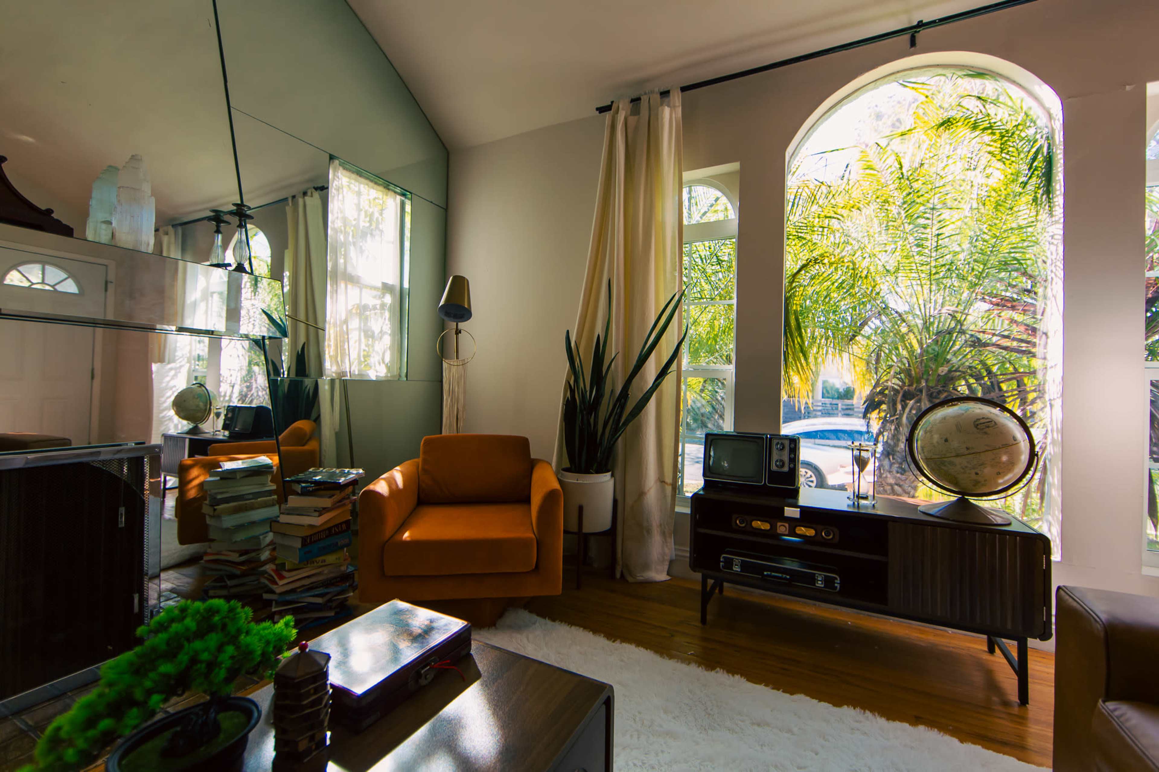 A modern living room features an orange armchair, a wooden TV stand with a vintage globe, and large windows offering natural light that illuminates potted plants.