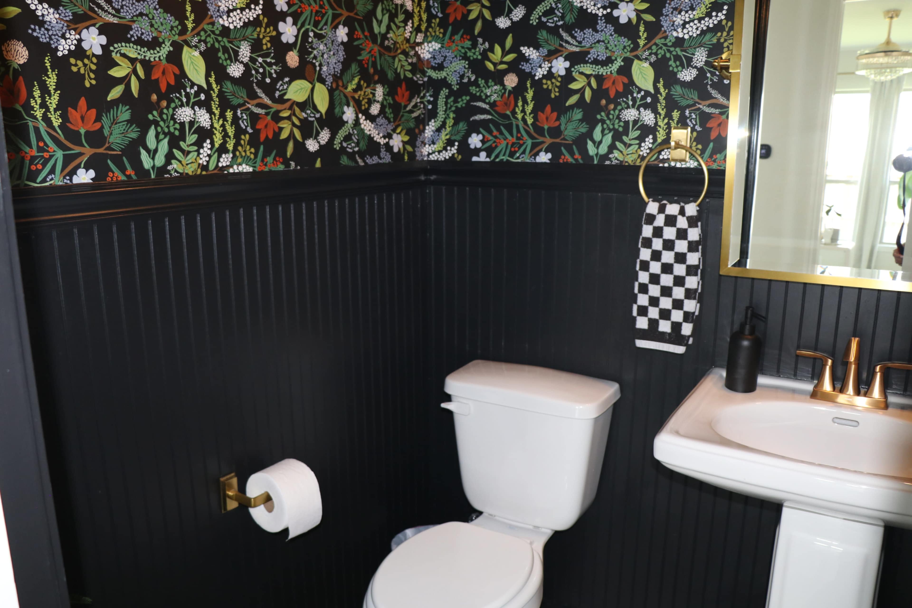 The image shows a small bathroom featuring a black wainscoting, floral wallpaper, a white toilet, sink, and a decorative towel holder.