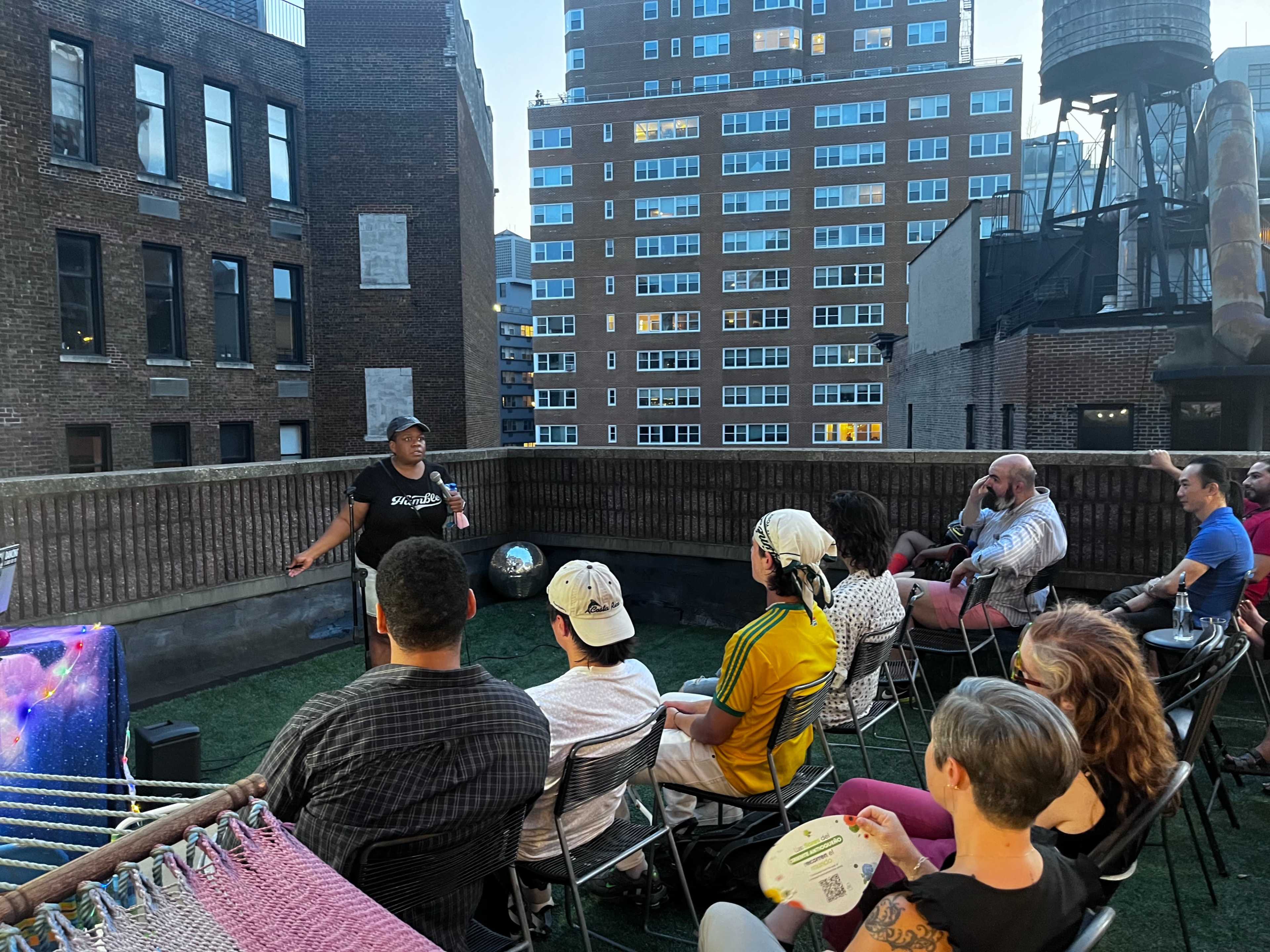 A speaker addresses an audience gathered on a rooftop with city buildings in the background.