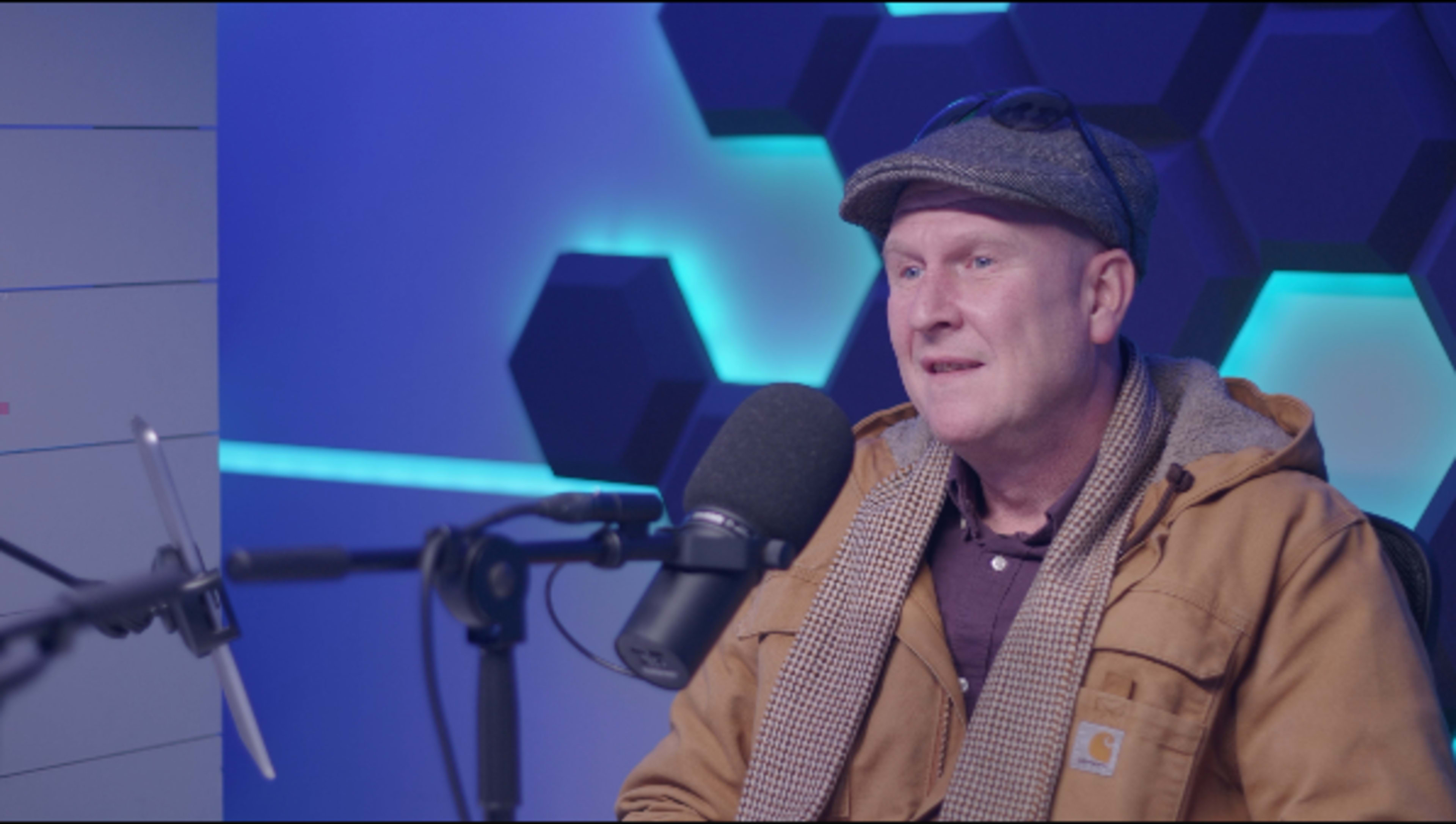 A man wearing a cap and scarf sits in front of a microphone in a studio with blue and hexagonal lighting.