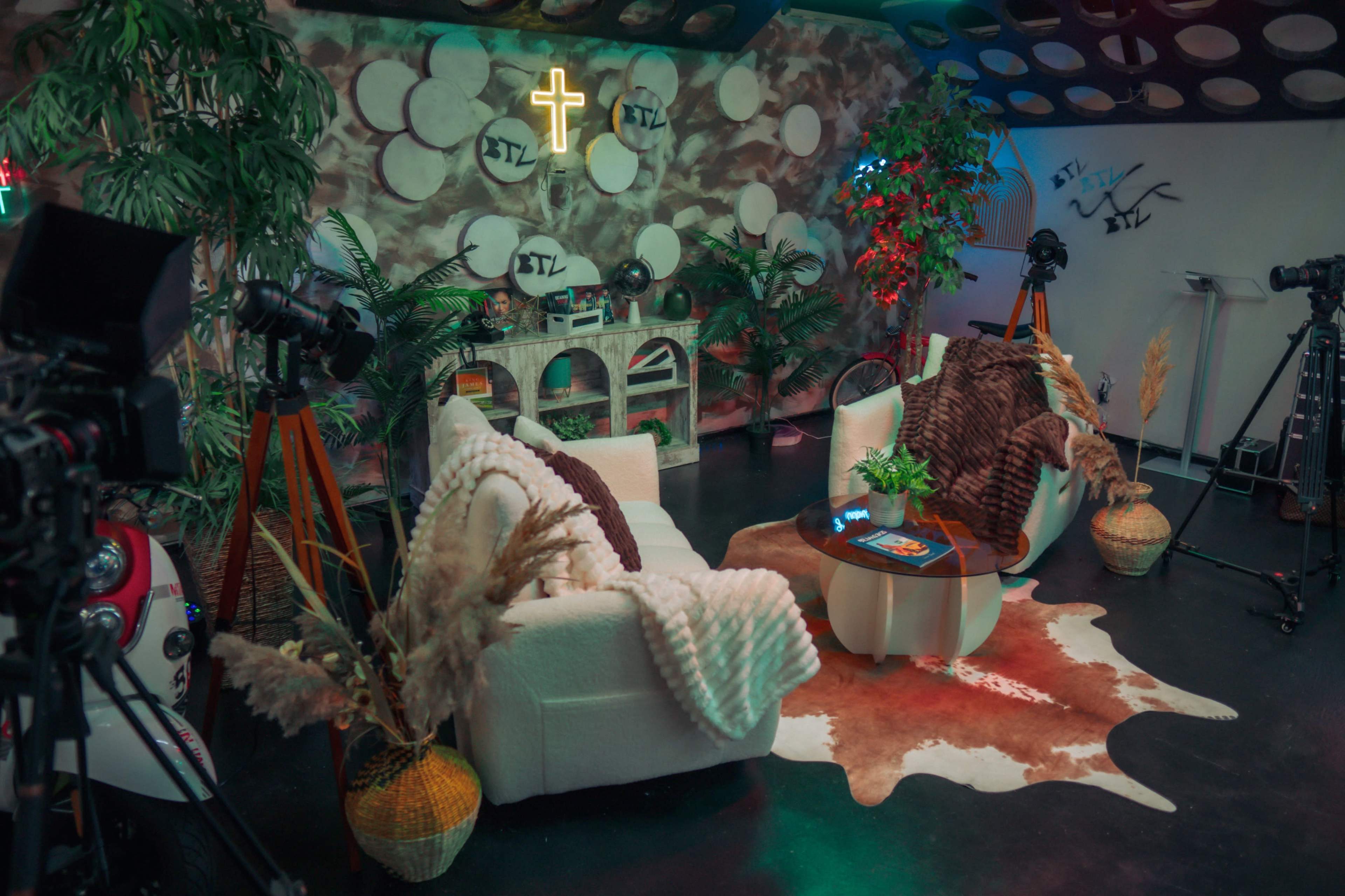The image shows a cozy studio setup with two white armchairs, a round coffee table on a patterned rug, surrounded by plants and cameras.