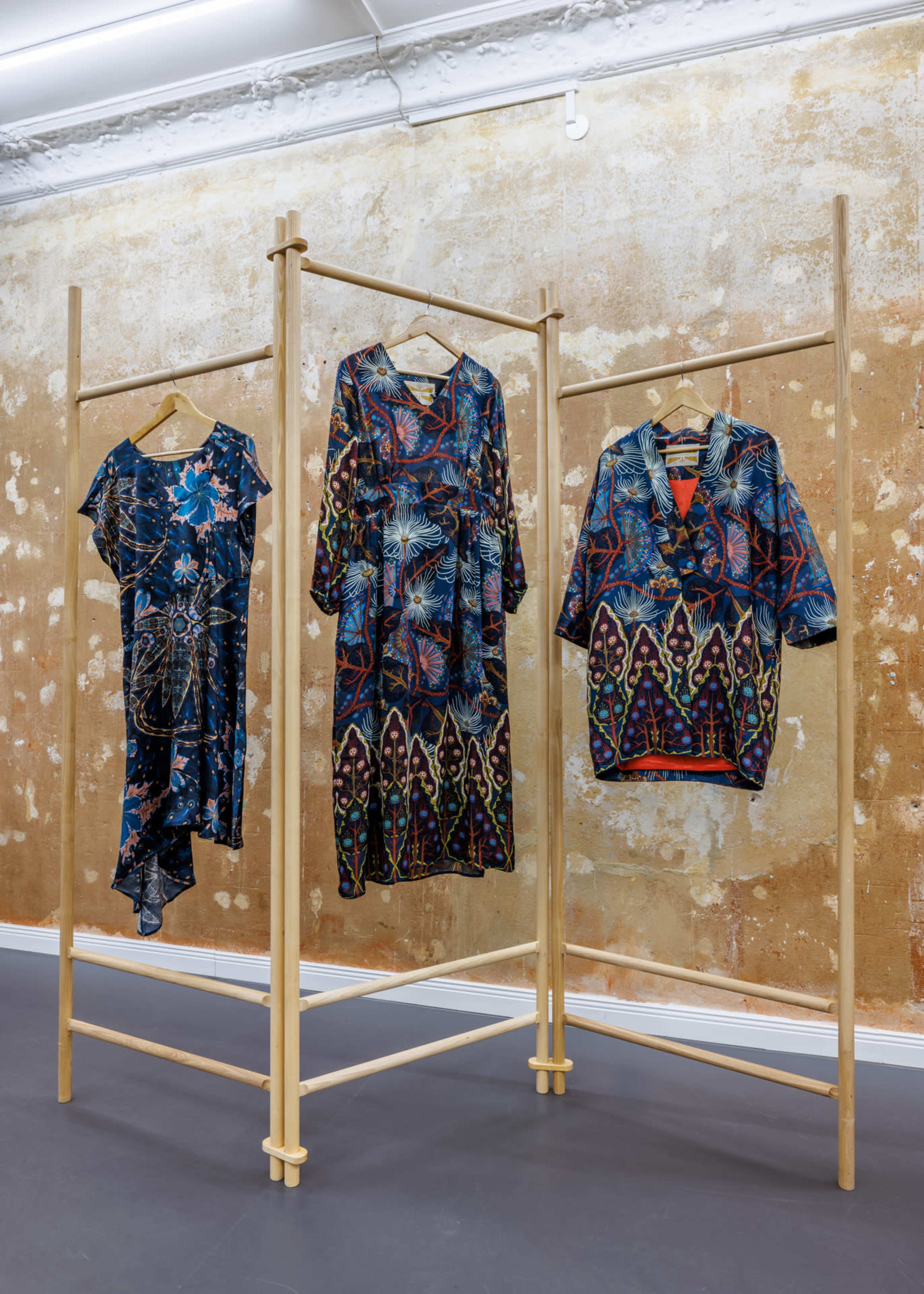 Three patterned dresses are displayed on wooden racks against a textured, rust-colored wall.