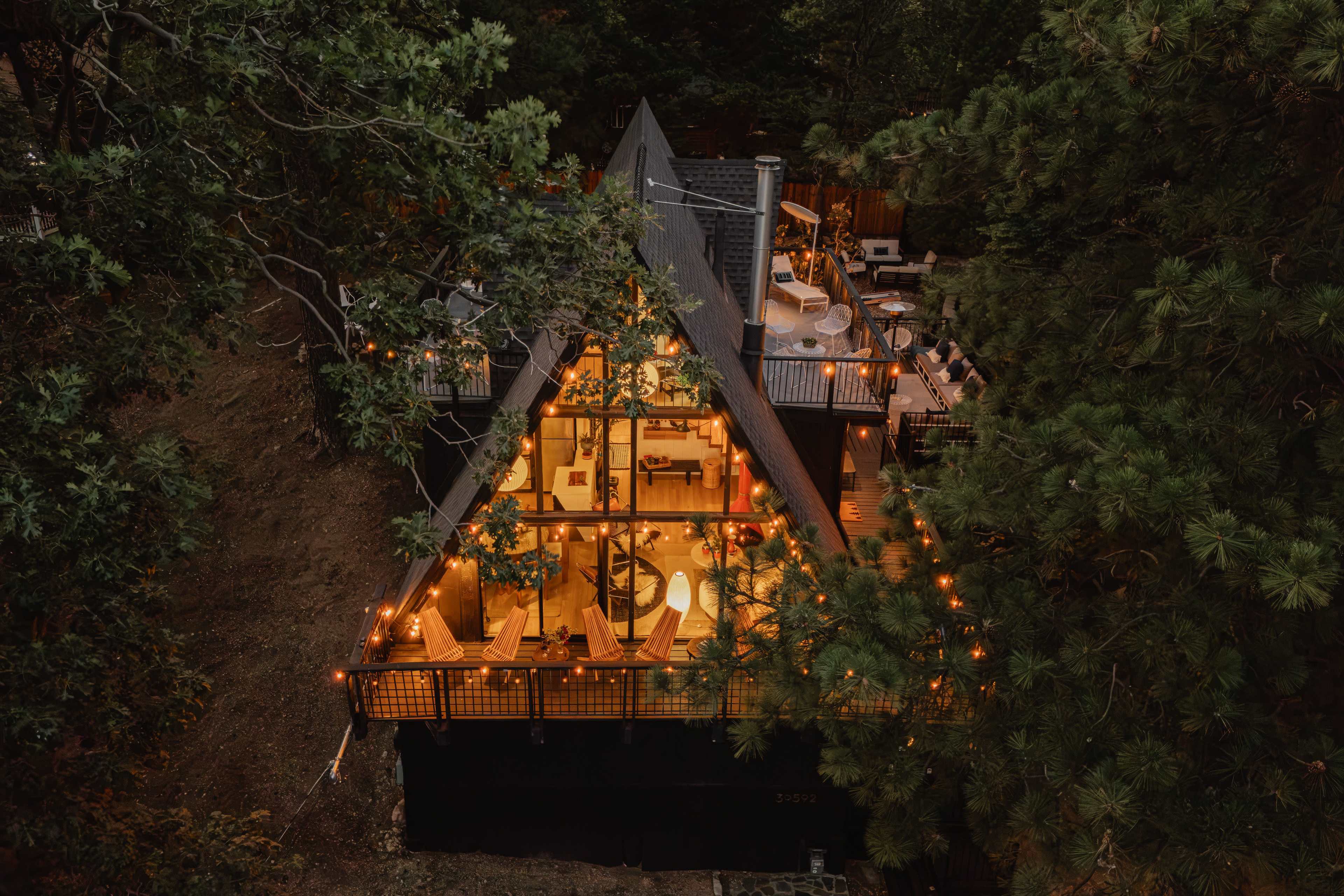 A modern, triangular-roofed house nestled among trees, illuminated by string lights, with outdoor seating areas visible on its deck.
