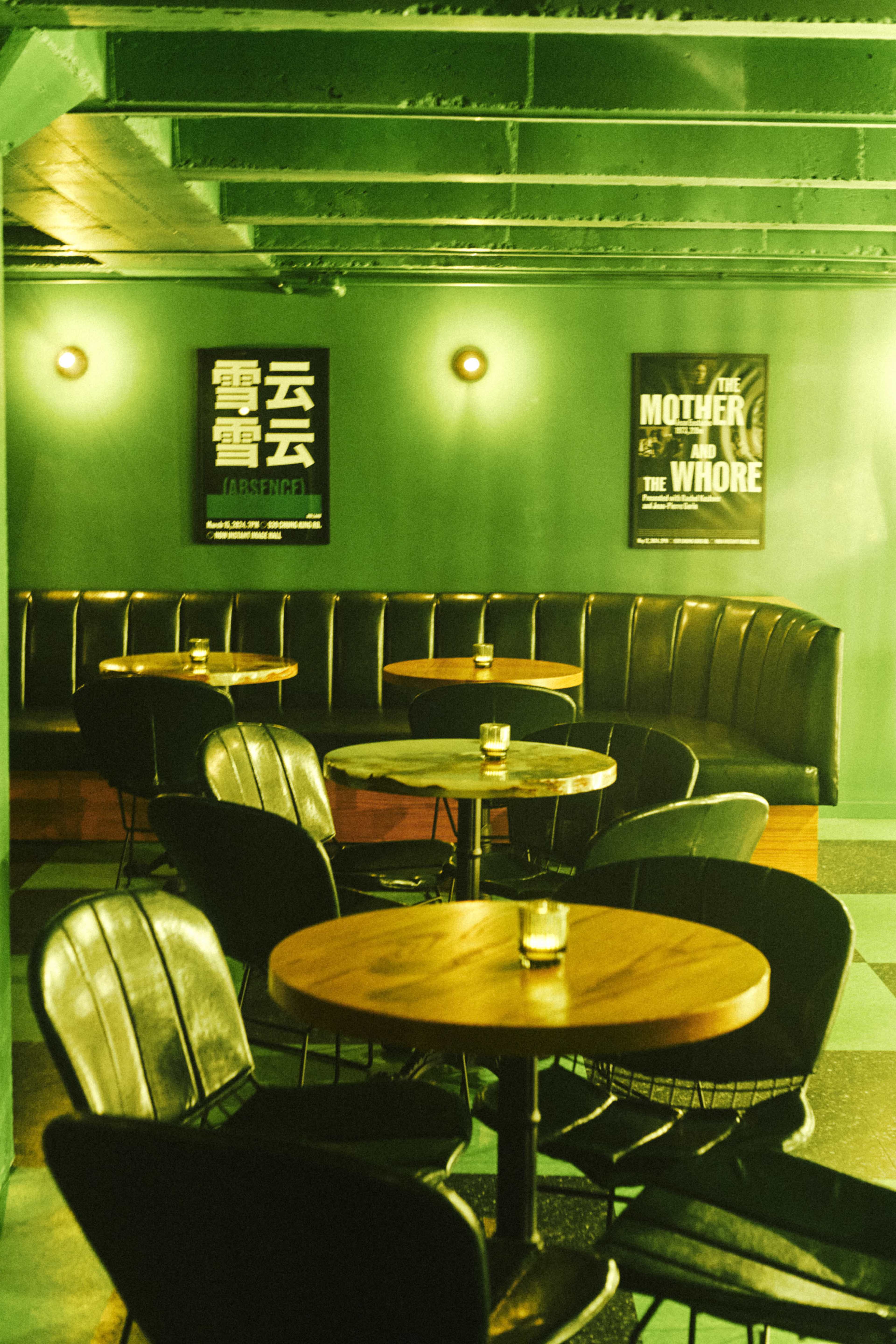 The image shows a dimly lit bar featuring black leather seating and round wooden tables, with posters hanging on the green walls.