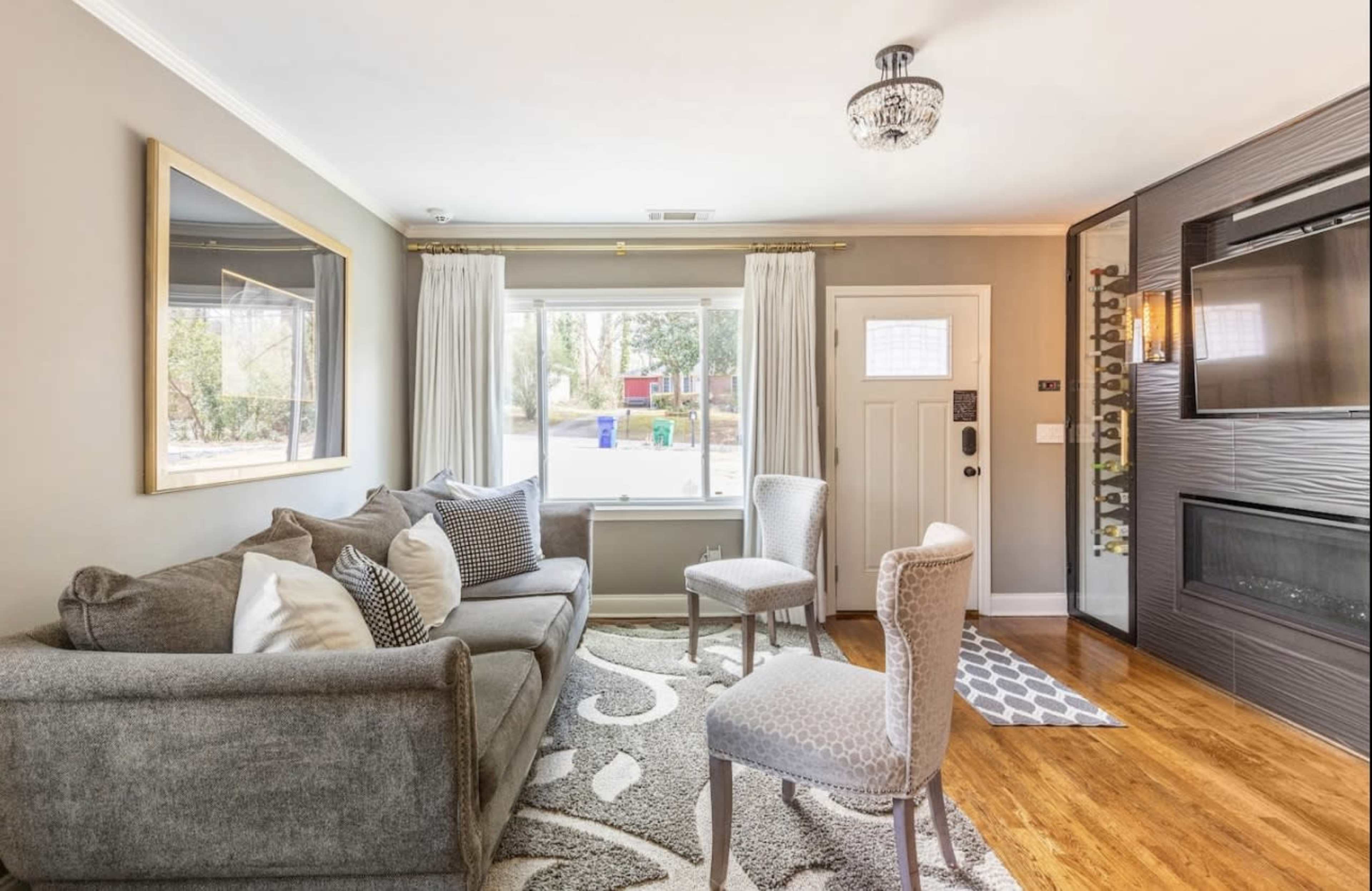 A modern living room with a gray sofa, two upholstered chairs, a patterned rug, and large windows facing a well-lit exterior.