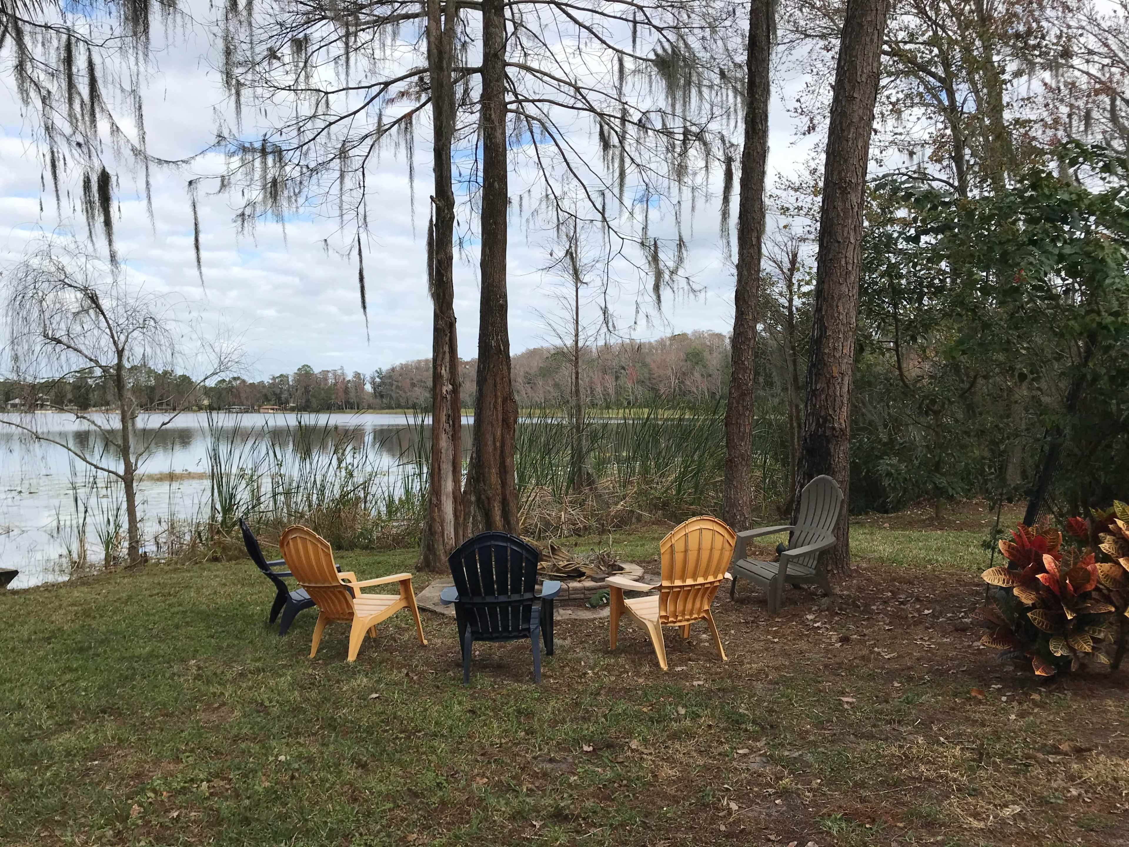 A circular arrangement of five outdoor chairs is situated near a calm lake surrounded by greenery and trees.