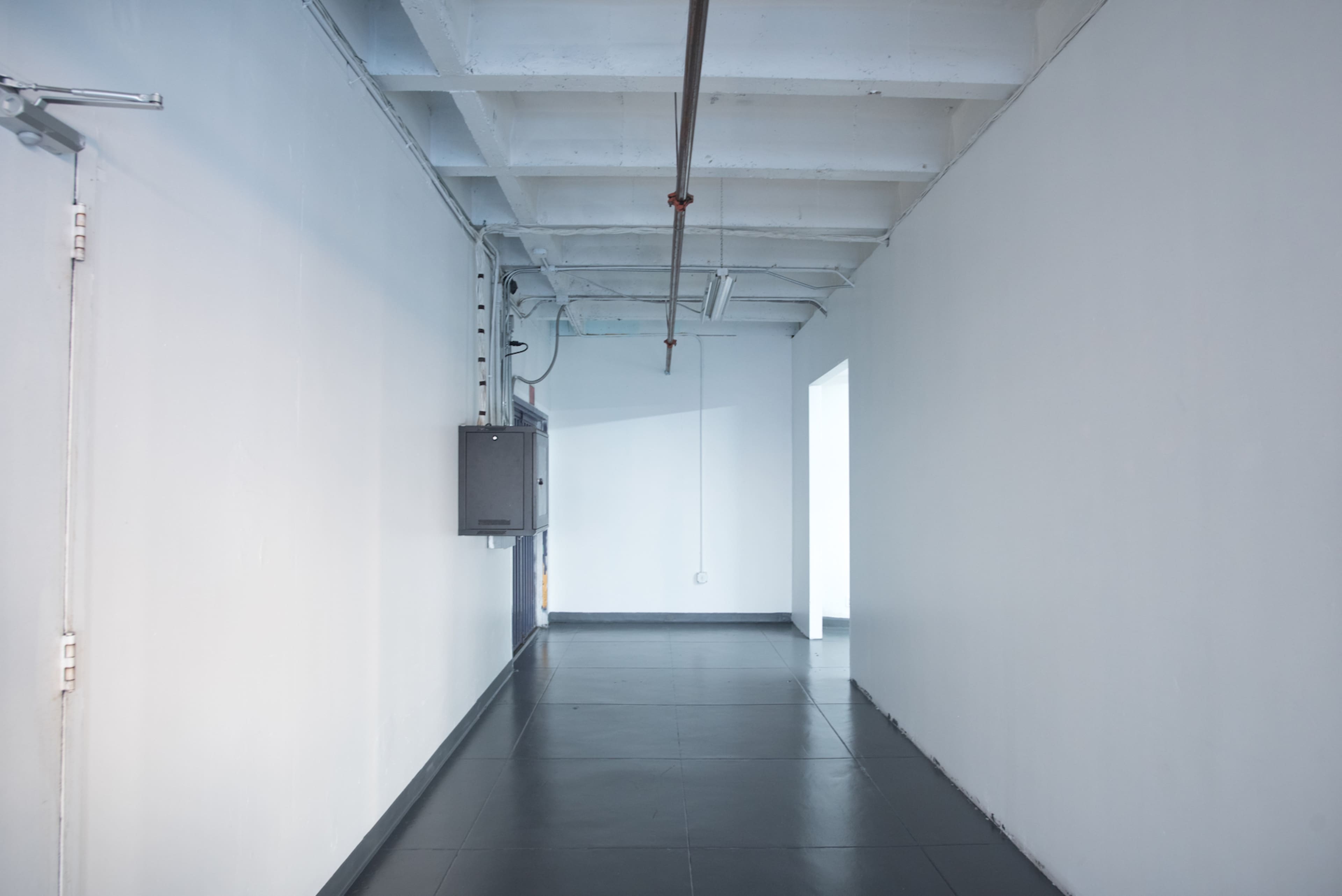 A narrow, empty hallway with white walls and a dark floor, featuring a small electrical box on the side.