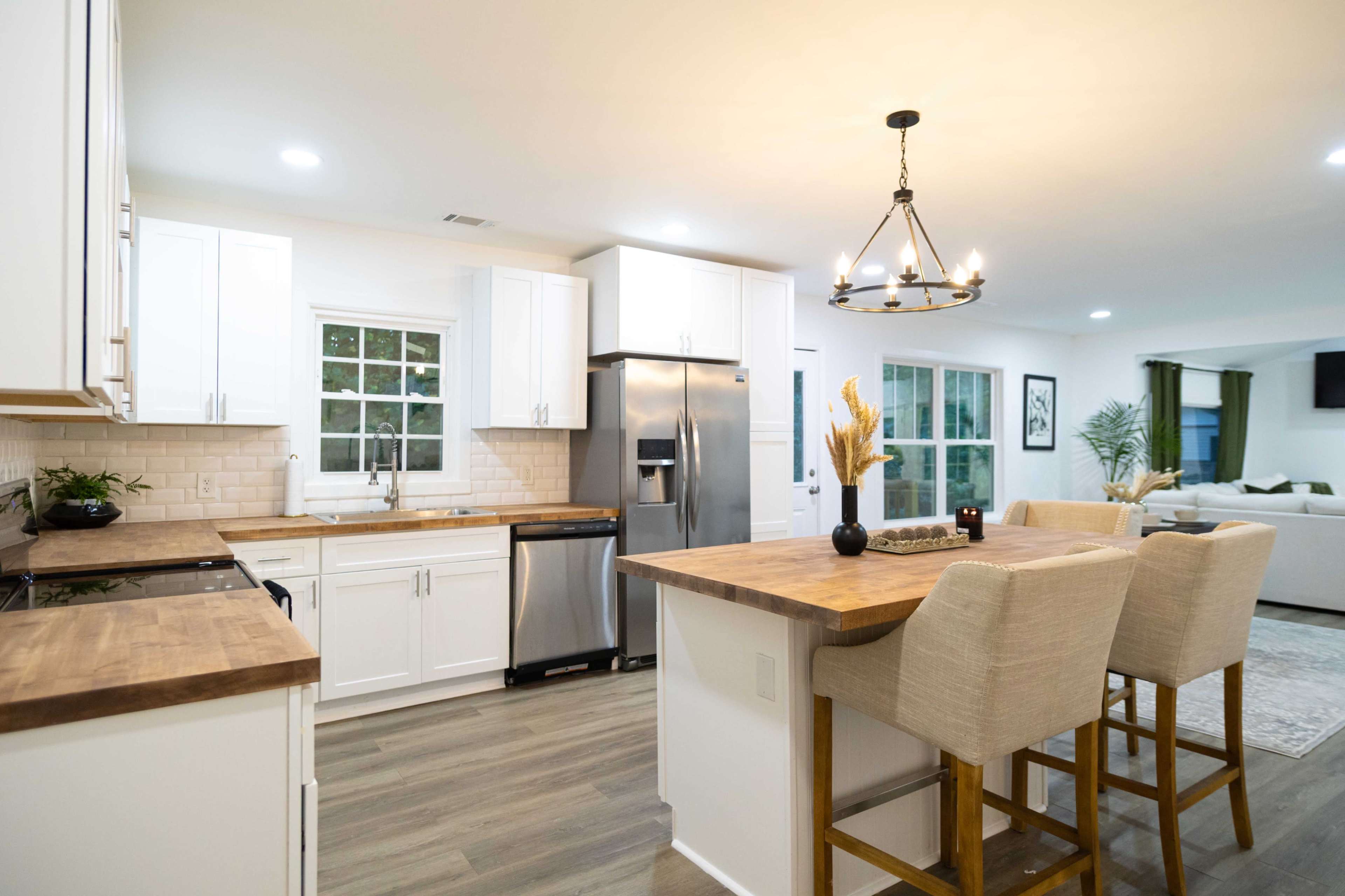 The image depicts a modern kitchen with white cabinetry, wooden countertops, stainless steel appliances, and a dining area featuring tall chairs and a decorative chandelier.