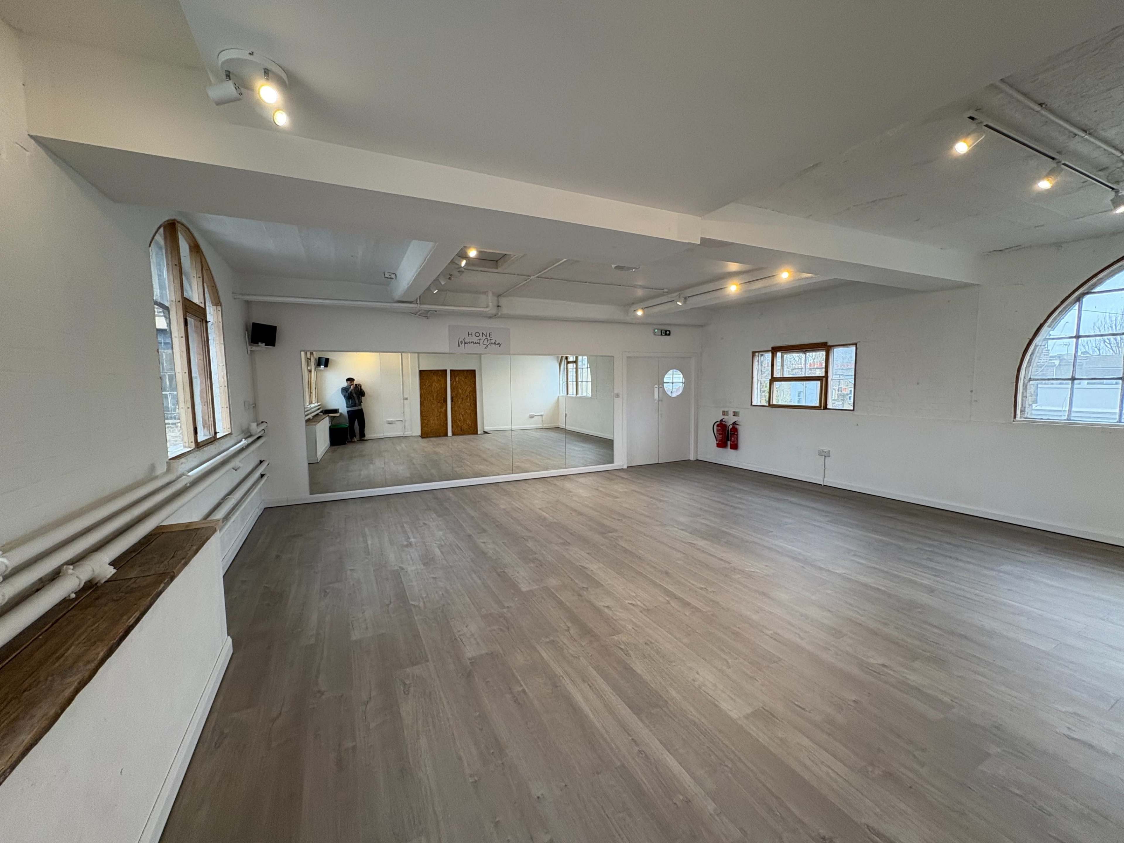 The image shows an empty studio space with wooden flooring, large windows, and a mirror on one wall.