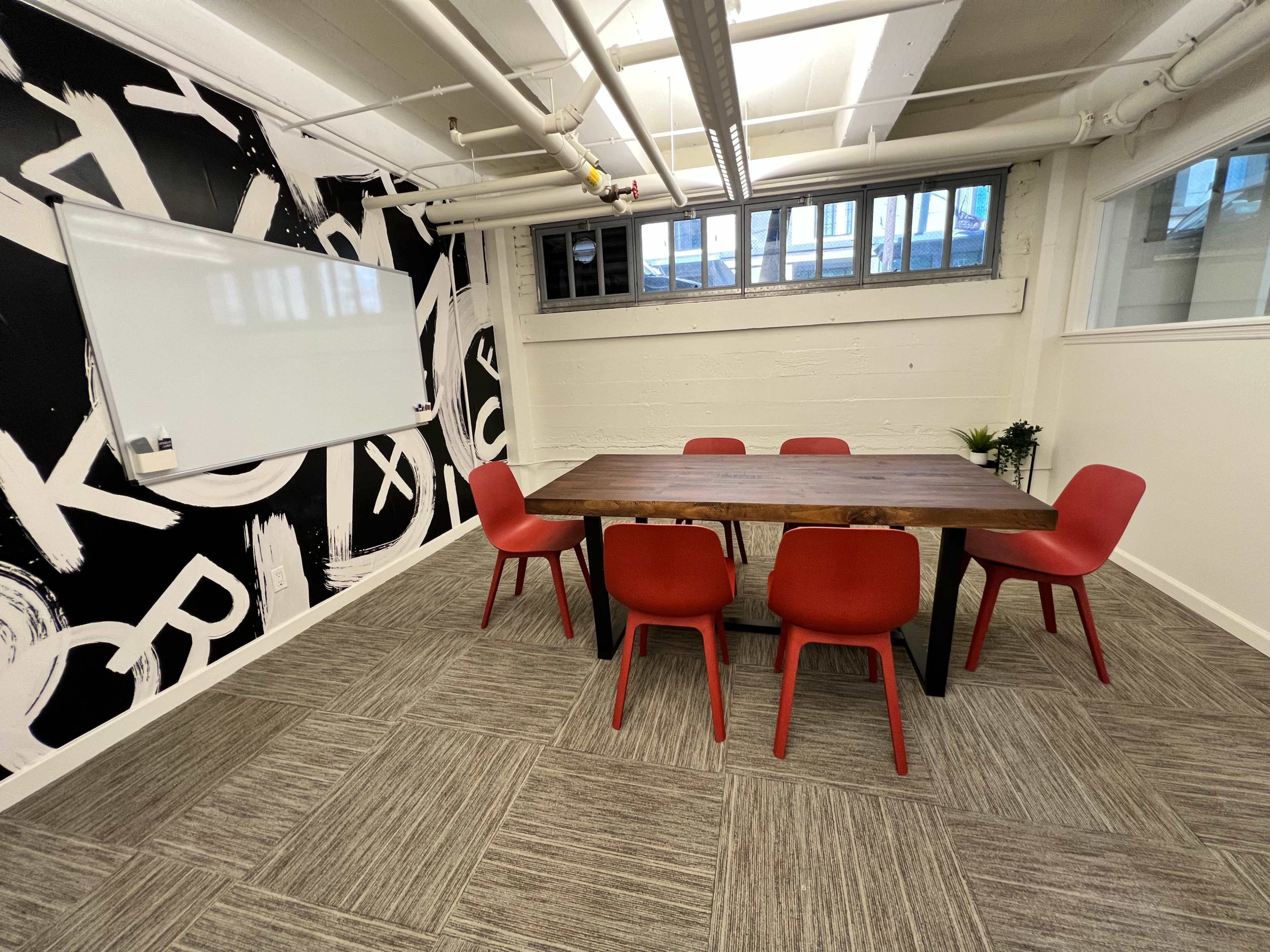 A conference room features a wooden table surrounded by six red chairs, with a whiteboard on one wall and a graphic black-and-white mural on another.