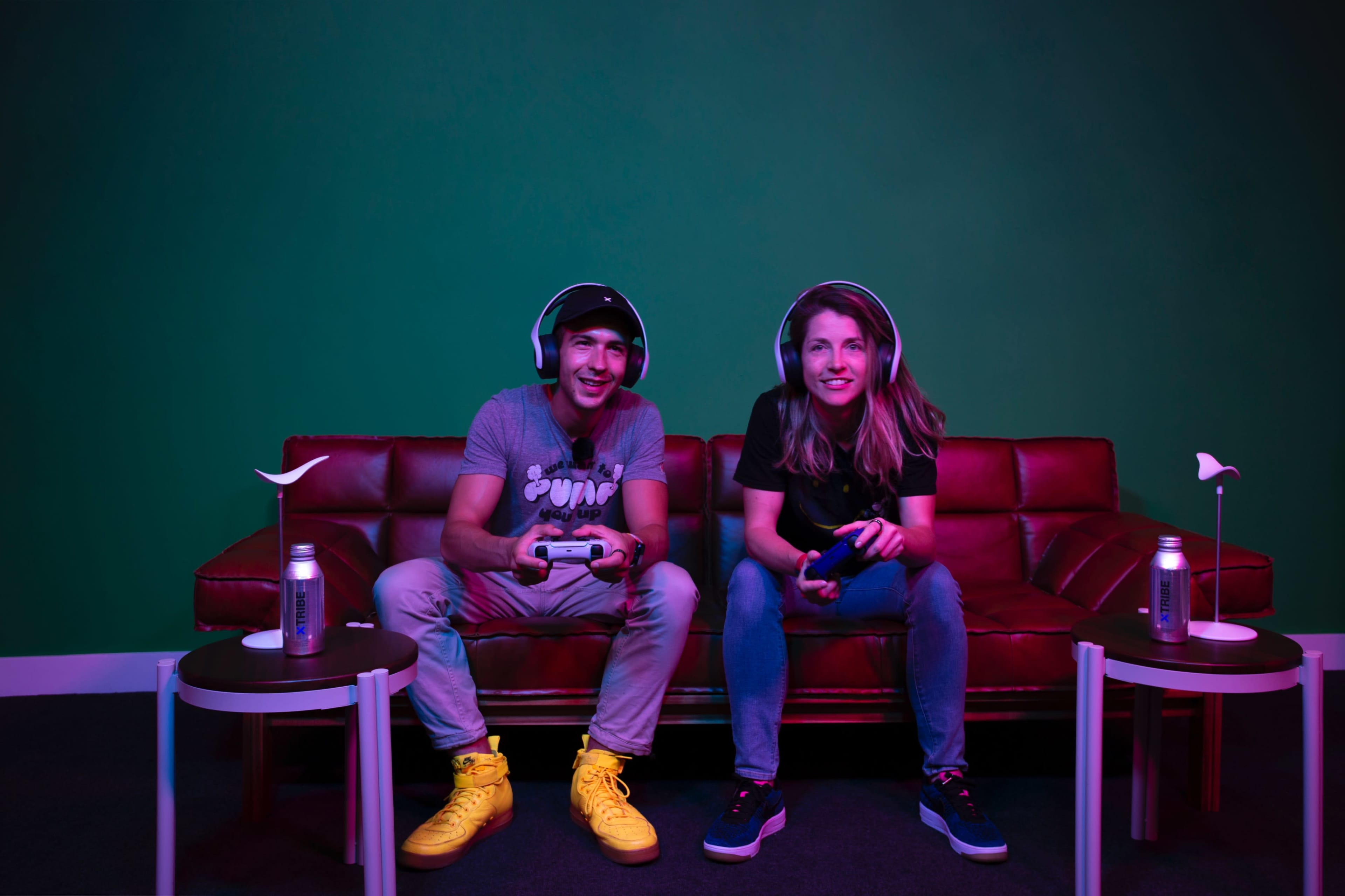 Two people sit on a red couch with gaming headsets, holding controllers while playing video games in a room with a green wall.