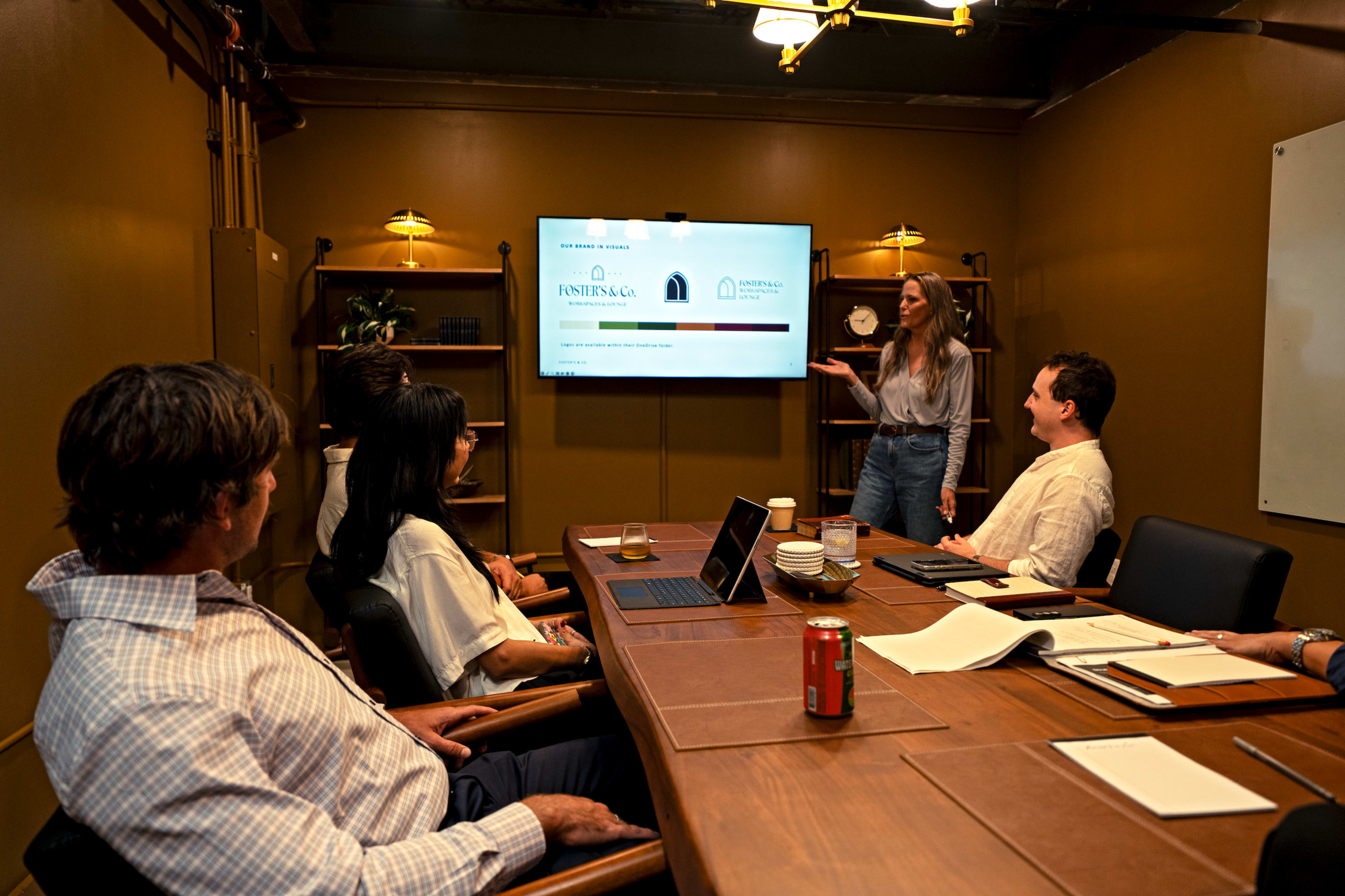A woman presents information on a screen to a group of four people seated around a table in a meeting room.