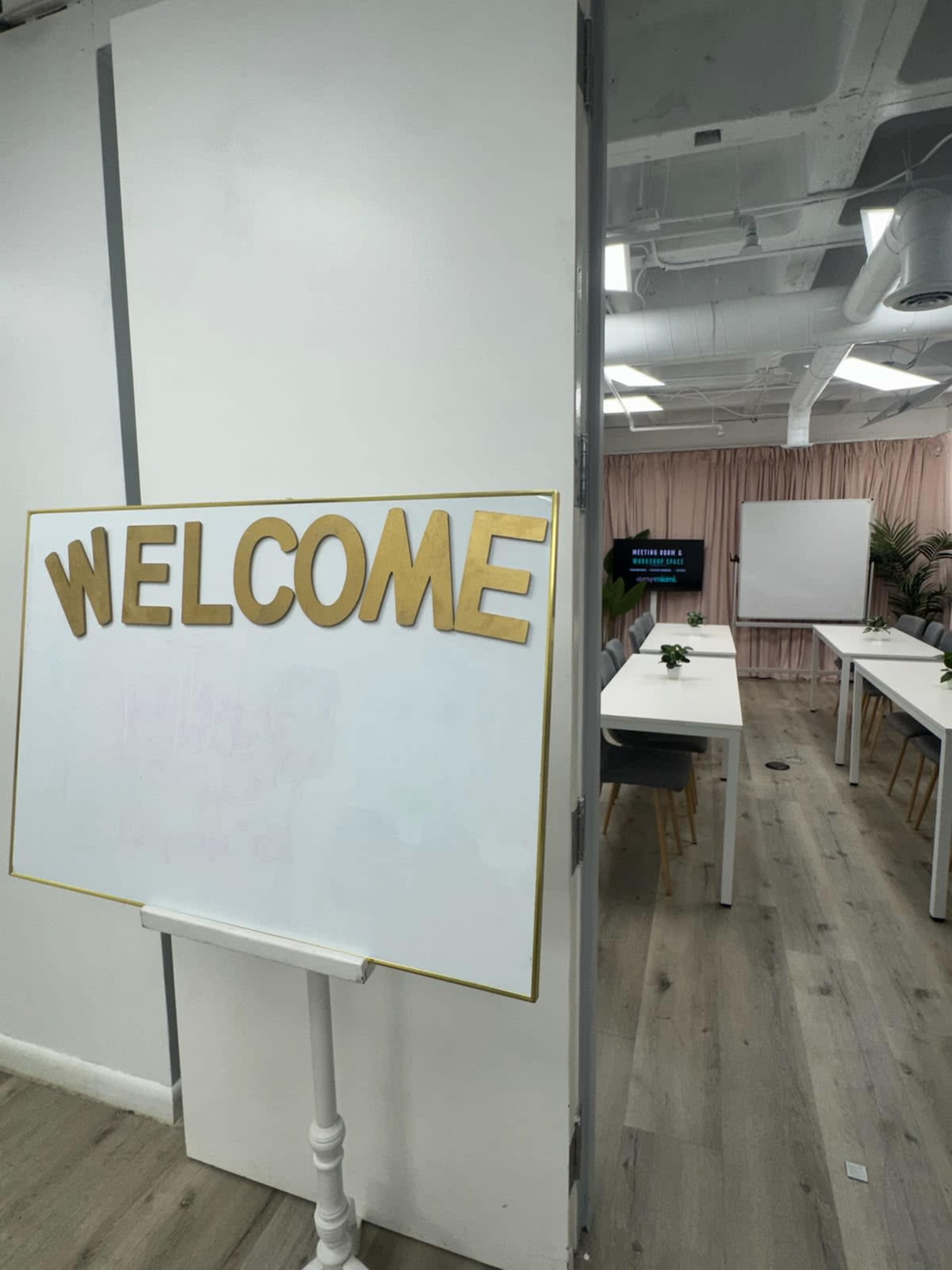 The image shows a brightly lit room with a large sign that says "WELCOME" on a whiteboard, accompanied by empty tables and a projector screen in the background.
