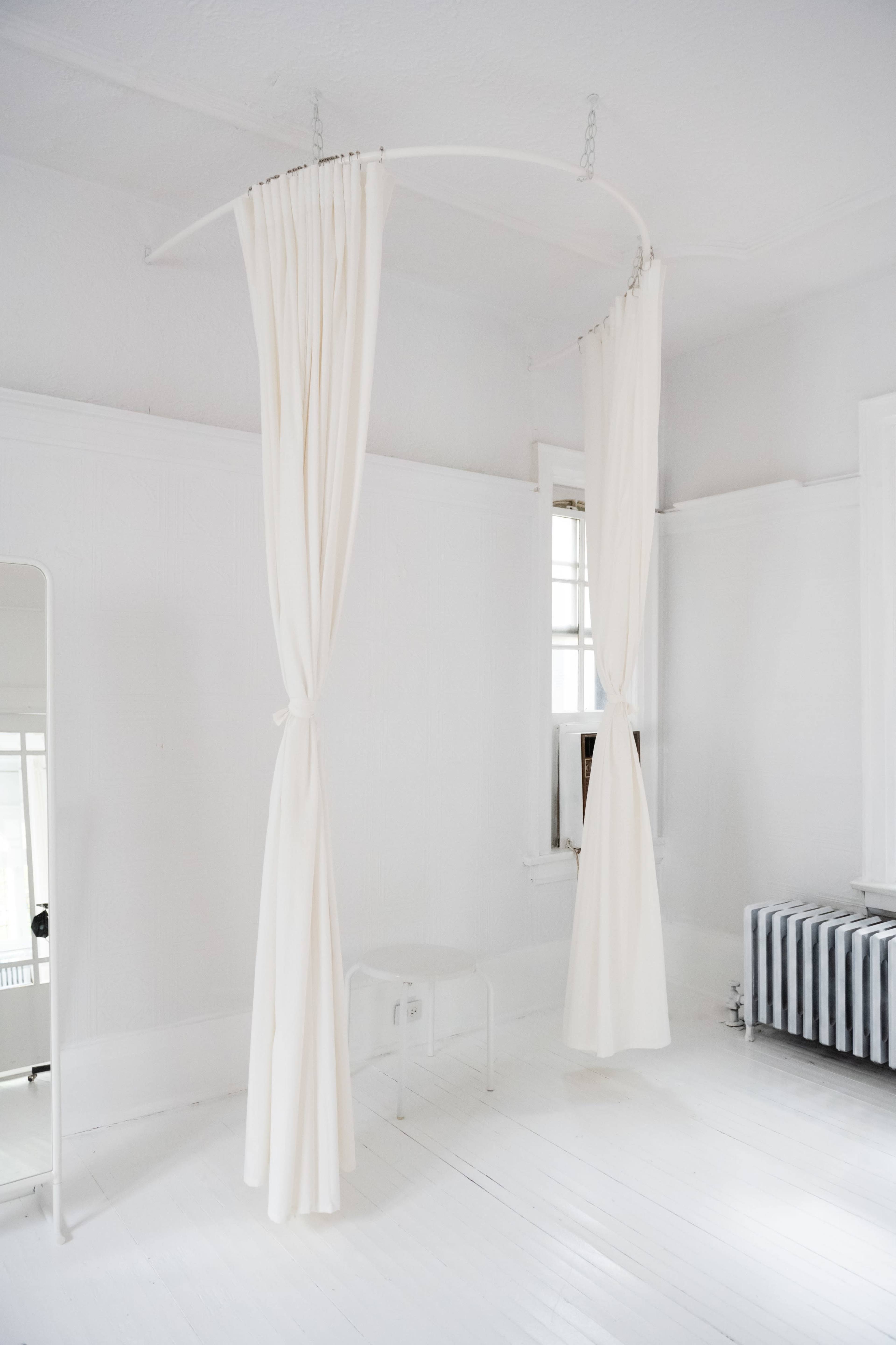 A bright, minimalist room with white walls and a wooden floor, featuring hanging white curtains on a curved rod and a striped radiator.